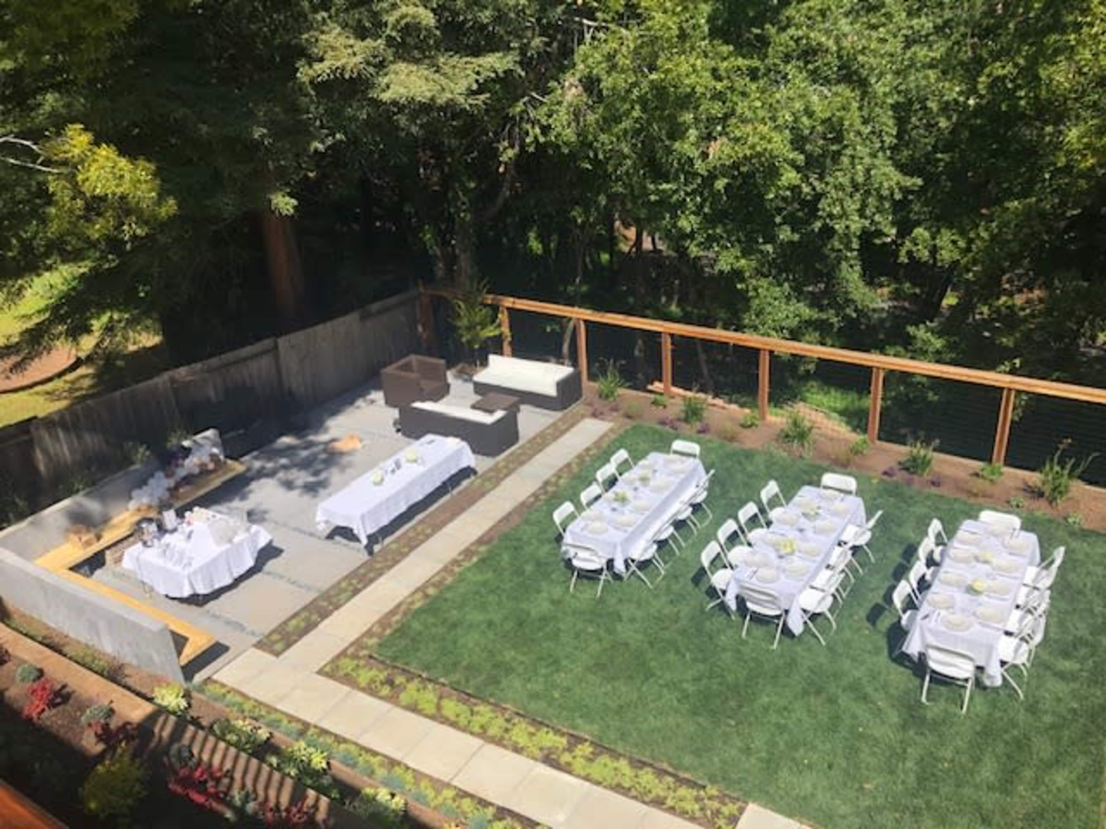 An outdoor event space with neatly arranged tables and chairs set on a lawn, surrounded by greenery and a wooden fence.