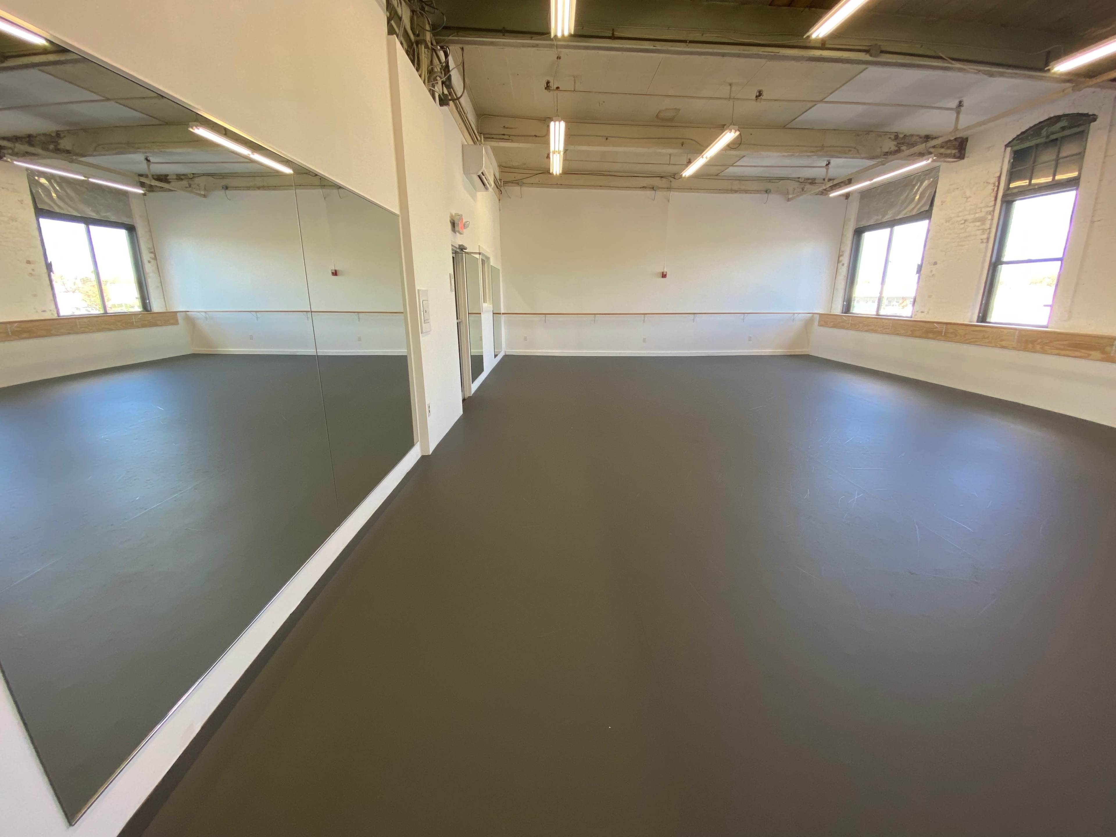 A spacious dance studio features a smooth, dark floor and large mirrors along one wall, with natural light coming through tall windows.