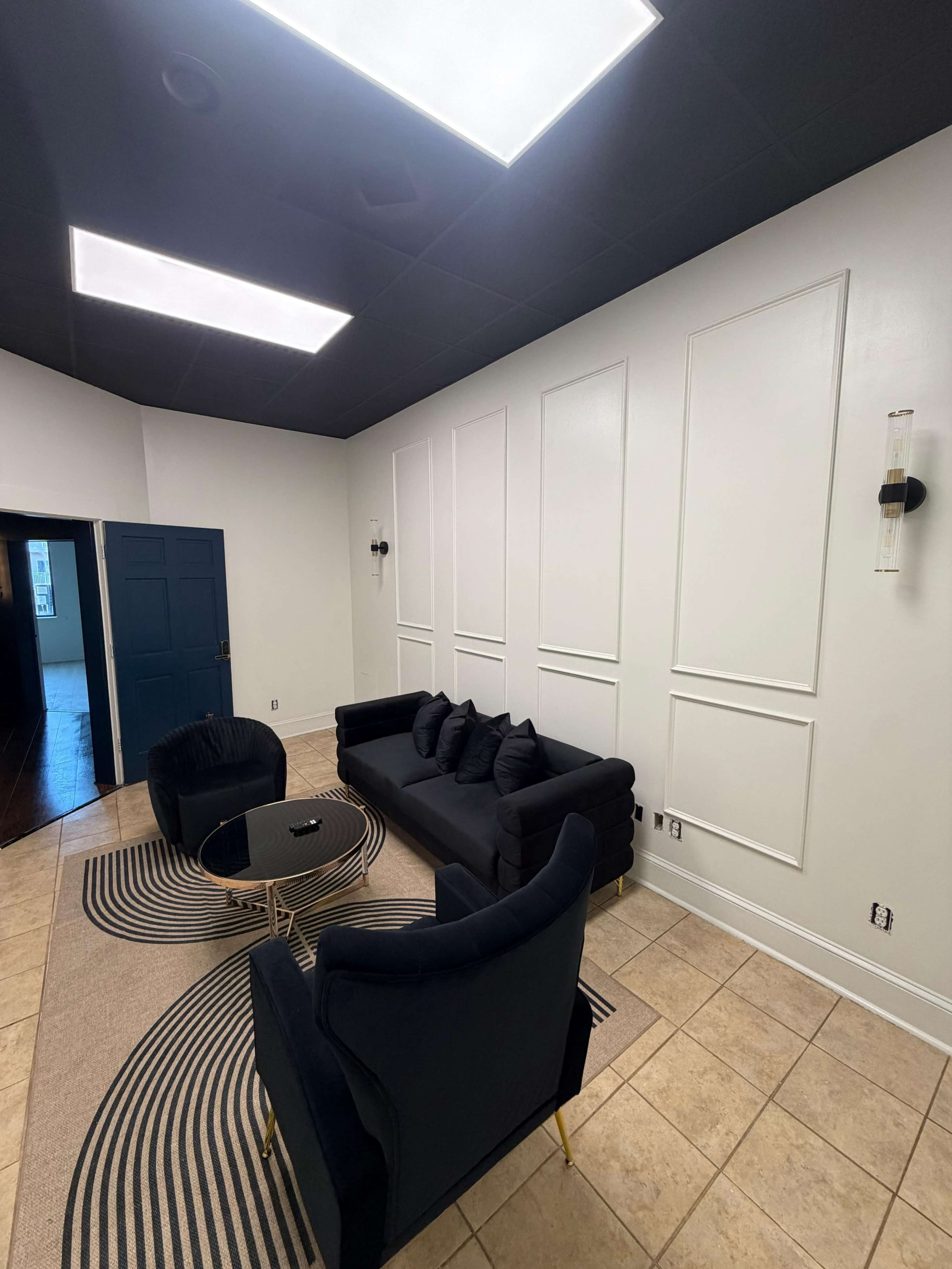 The image shows a modern lounge area featuring a dark sectional sofa, a round coffee table, and two matching chairs on a patterned rug, situated under a well-lit ceiling.