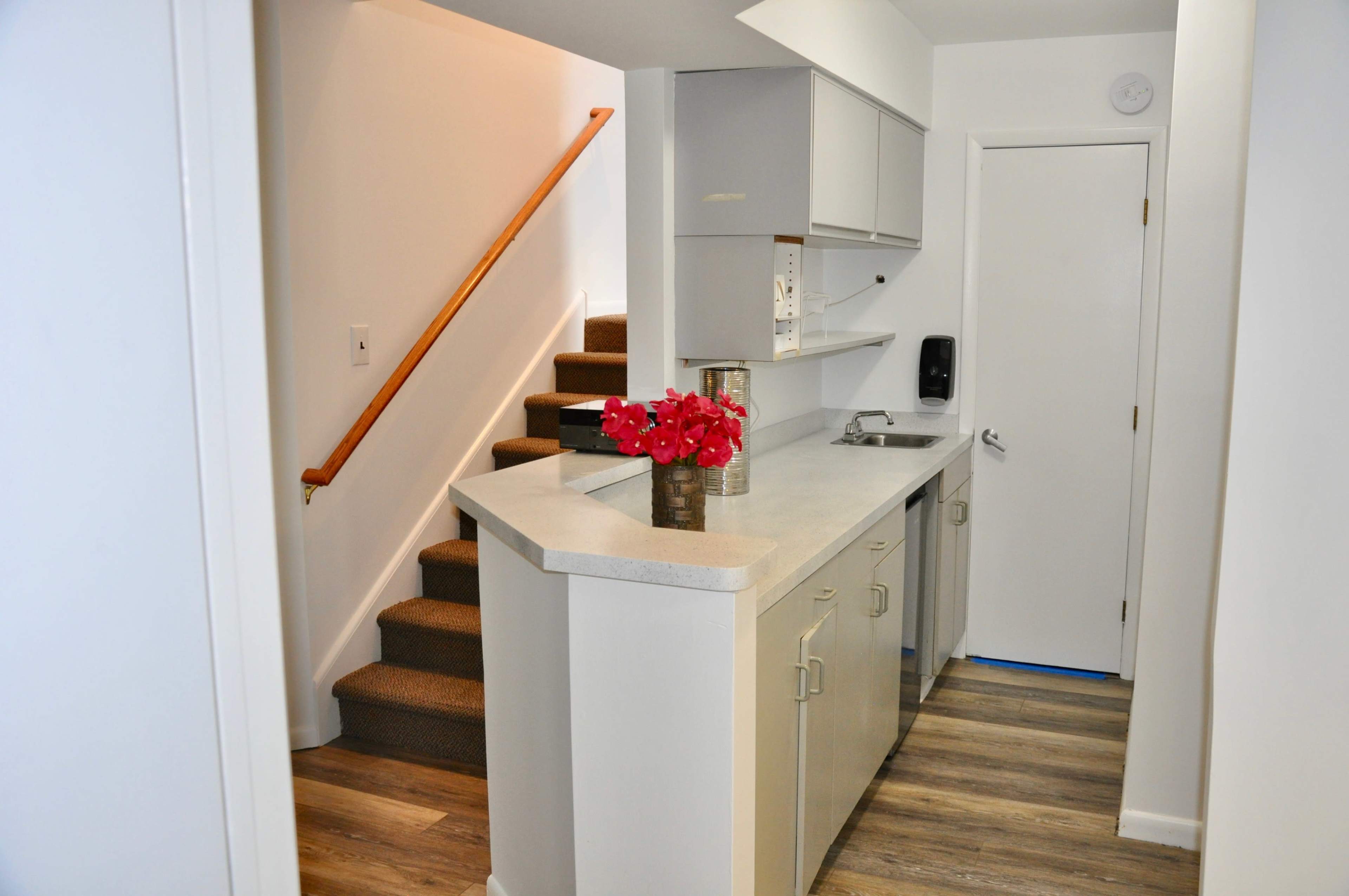 The image shows a modern kitchen area with a small island, a staircase in the background, and a white door leading to another room.