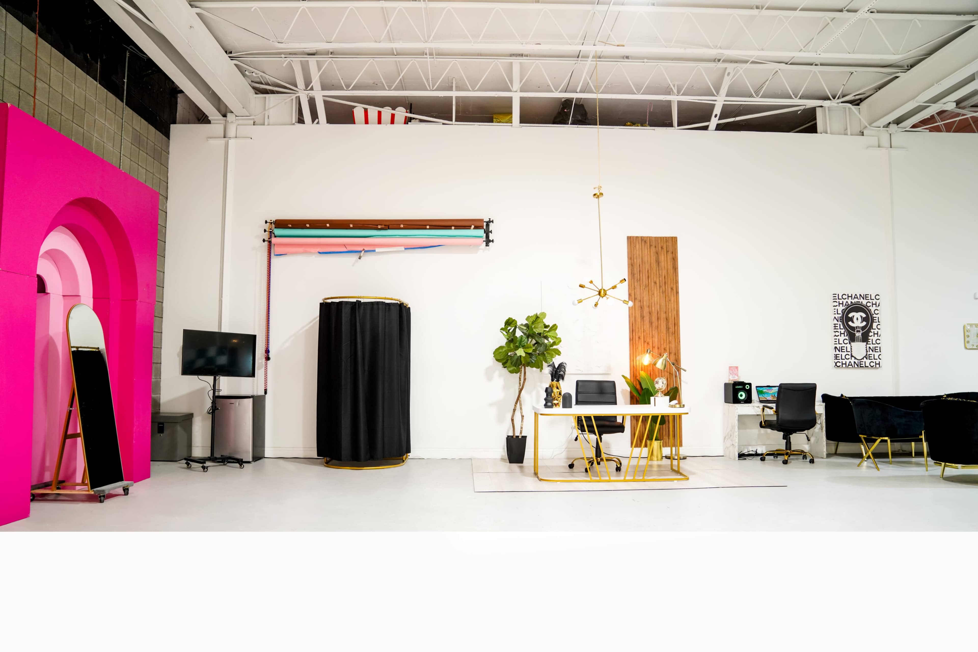 A modern, brightly lit studio space featuring a large pink wall, a table with decorative plants and equipment, and a backdrop setup for photography.