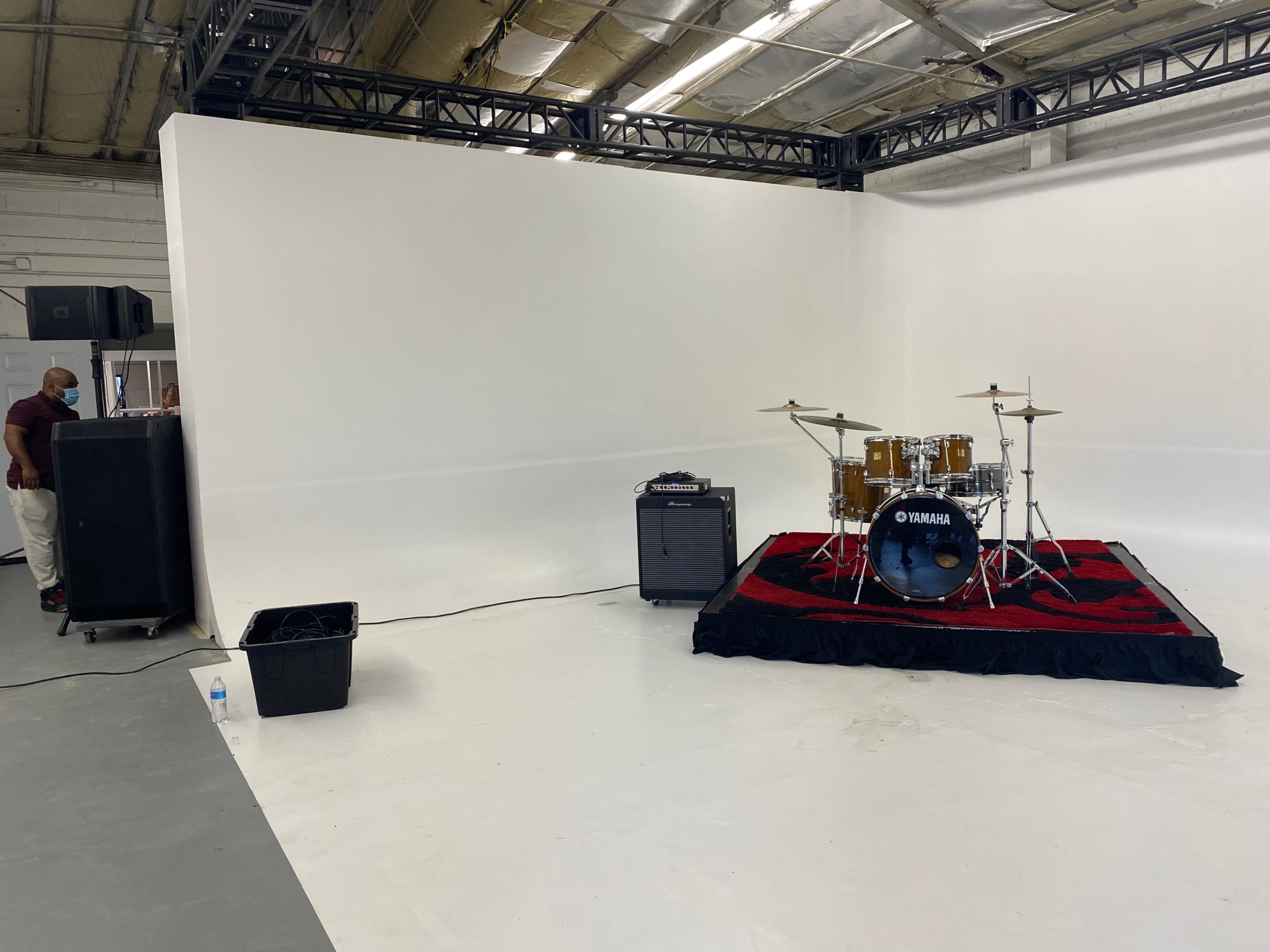A vacant studio space with a drum set on a small stage surrounded by a black amplifier and a plastic bin.