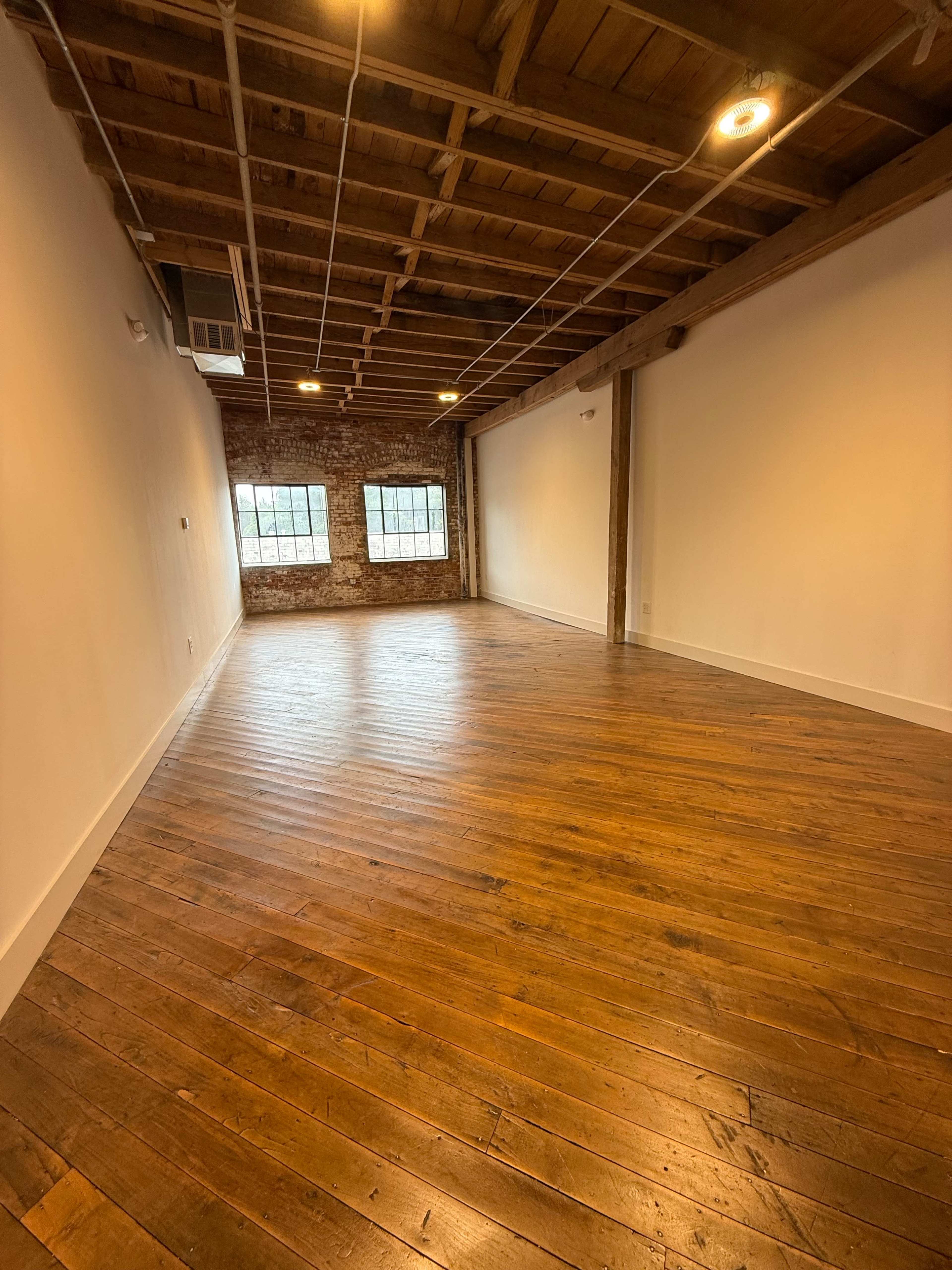 The image shows a spacious, empty room with wooden floors and exposed beams, featuring two large windows along one wall.