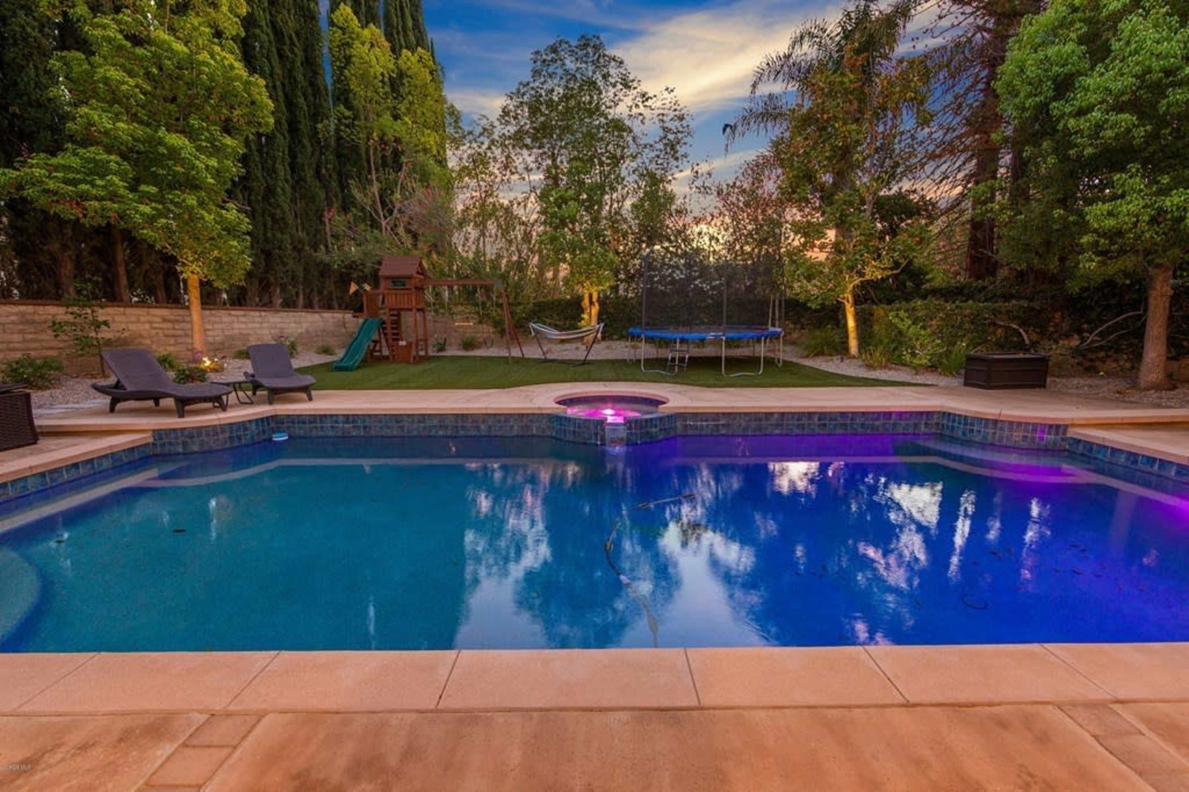The image shows a backyard pool area with lounge chairs, a jacuzzi, a trampoline, and a green lawn surrounded by trees.