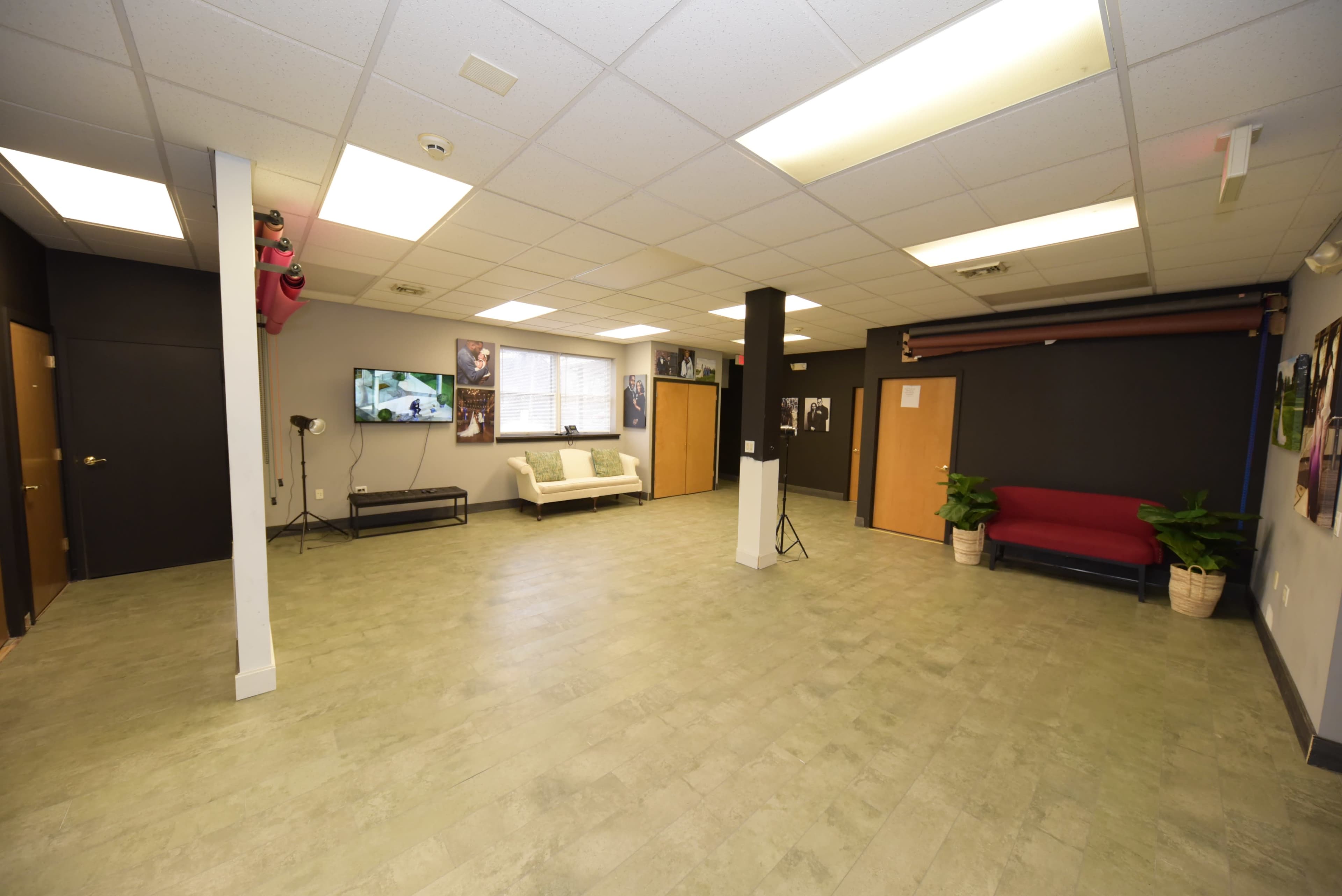 A spacious, minimally furnished room with a light-colored floor, black and beige walls, and several doors, featuring a couch, a red chair, and a television.