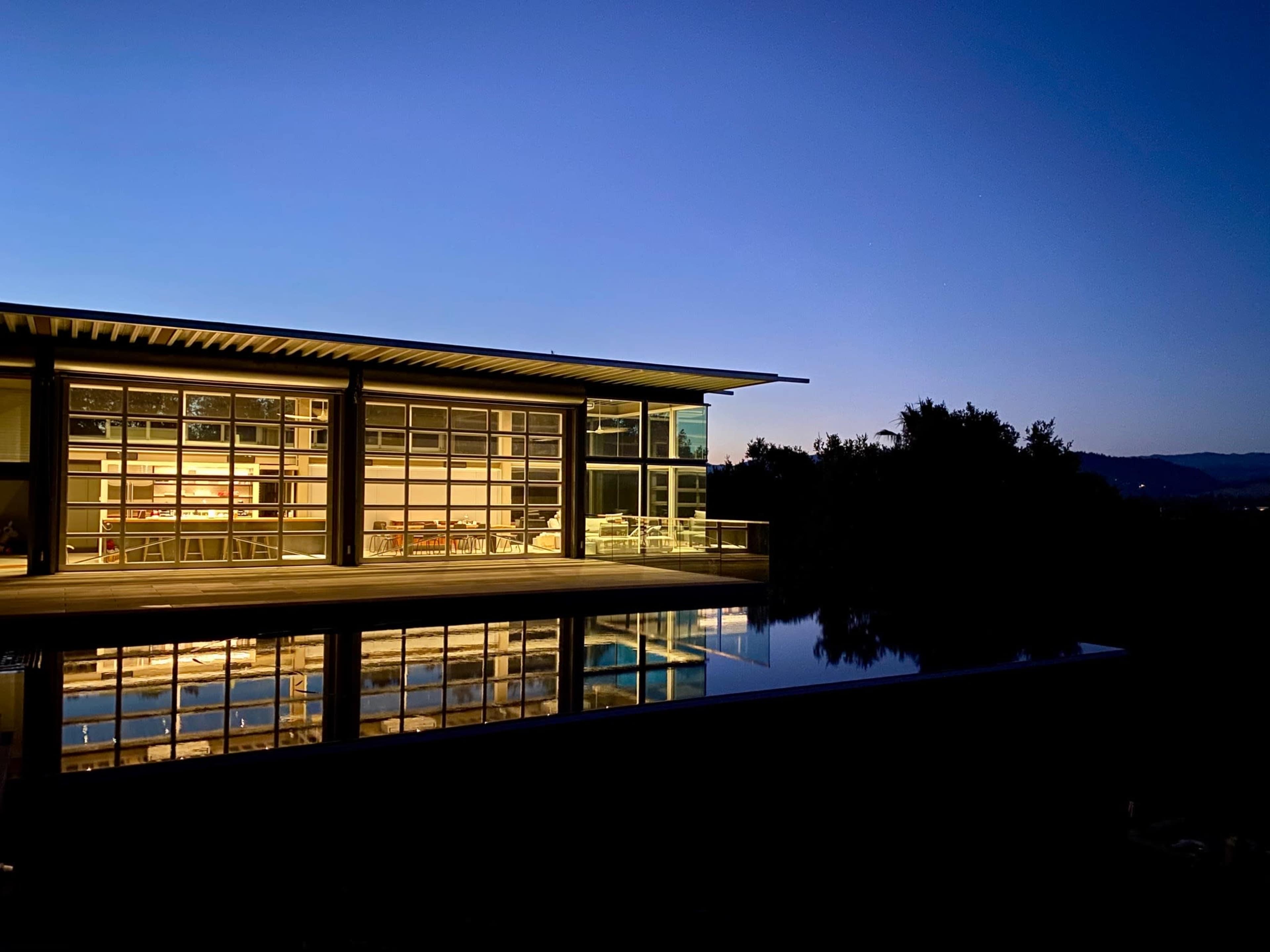 A modern building with large glass windows reflects in a still body of water under a twilight sky.