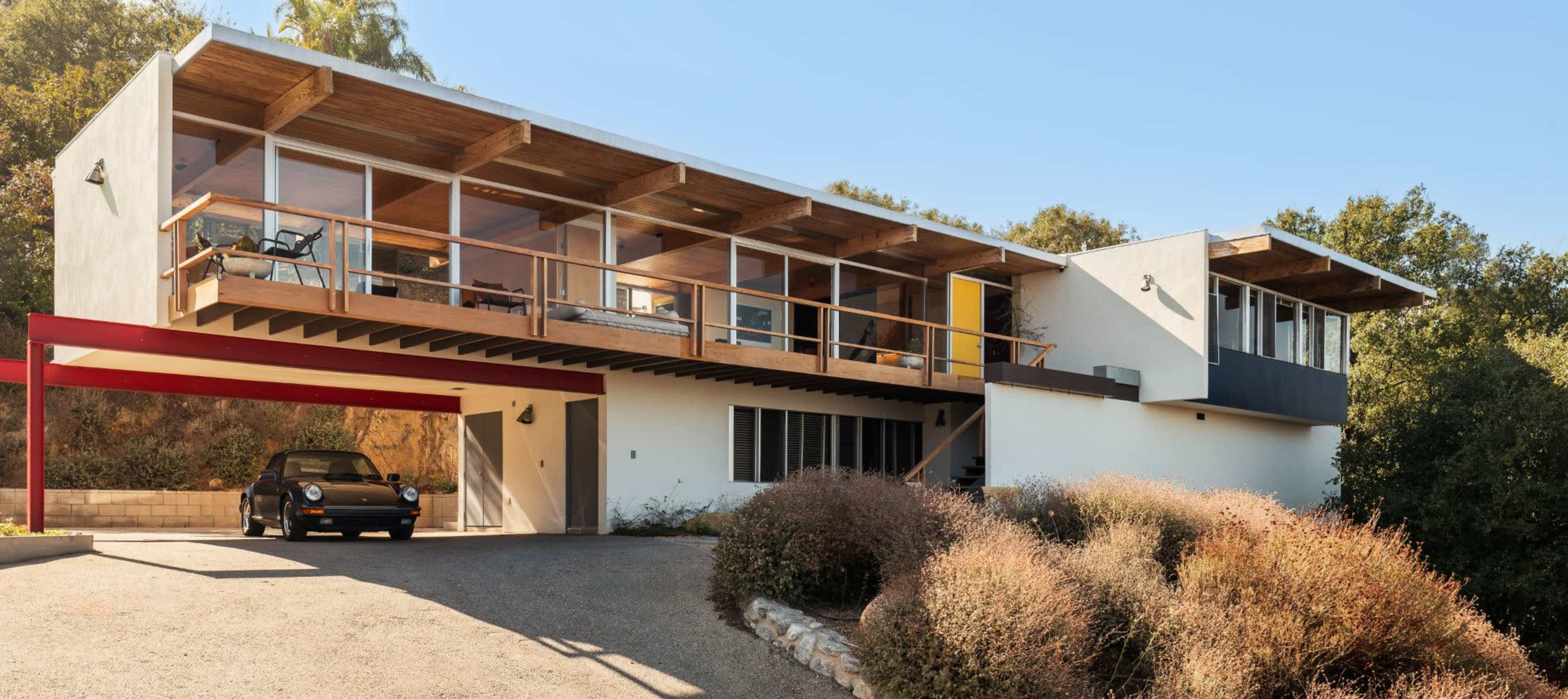 A modern house with large windows and a wooden deck is elevated on a slope, with a car parked in front under a carport.