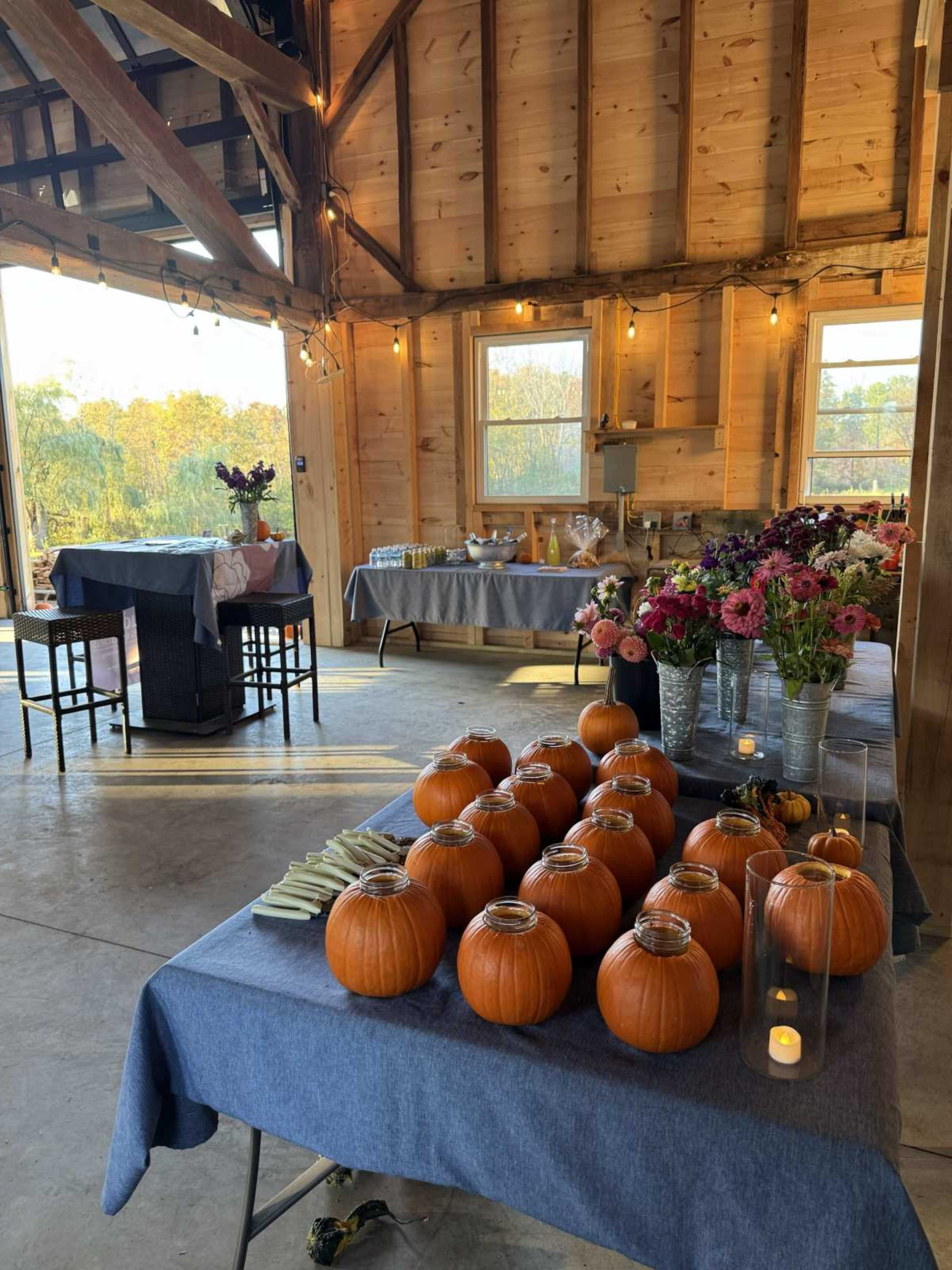 The image shows a rustic interior of a barn decorated with tables set for an event, featuring pumpkins and floral arrangements.