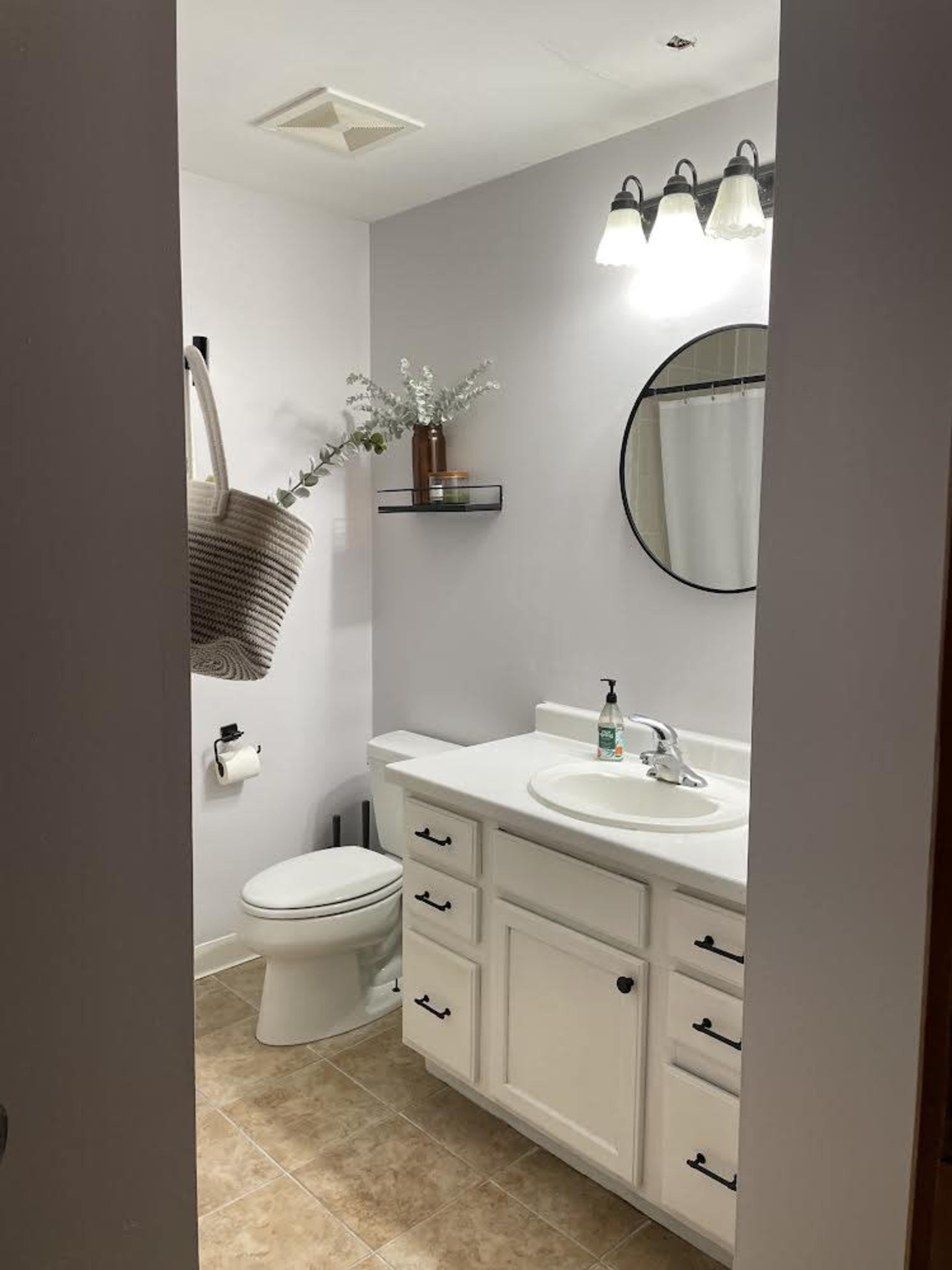 A bathroom featuring a toilet, sink with a mirror, and shelf, all against a light-colored wall and tiled floor.
