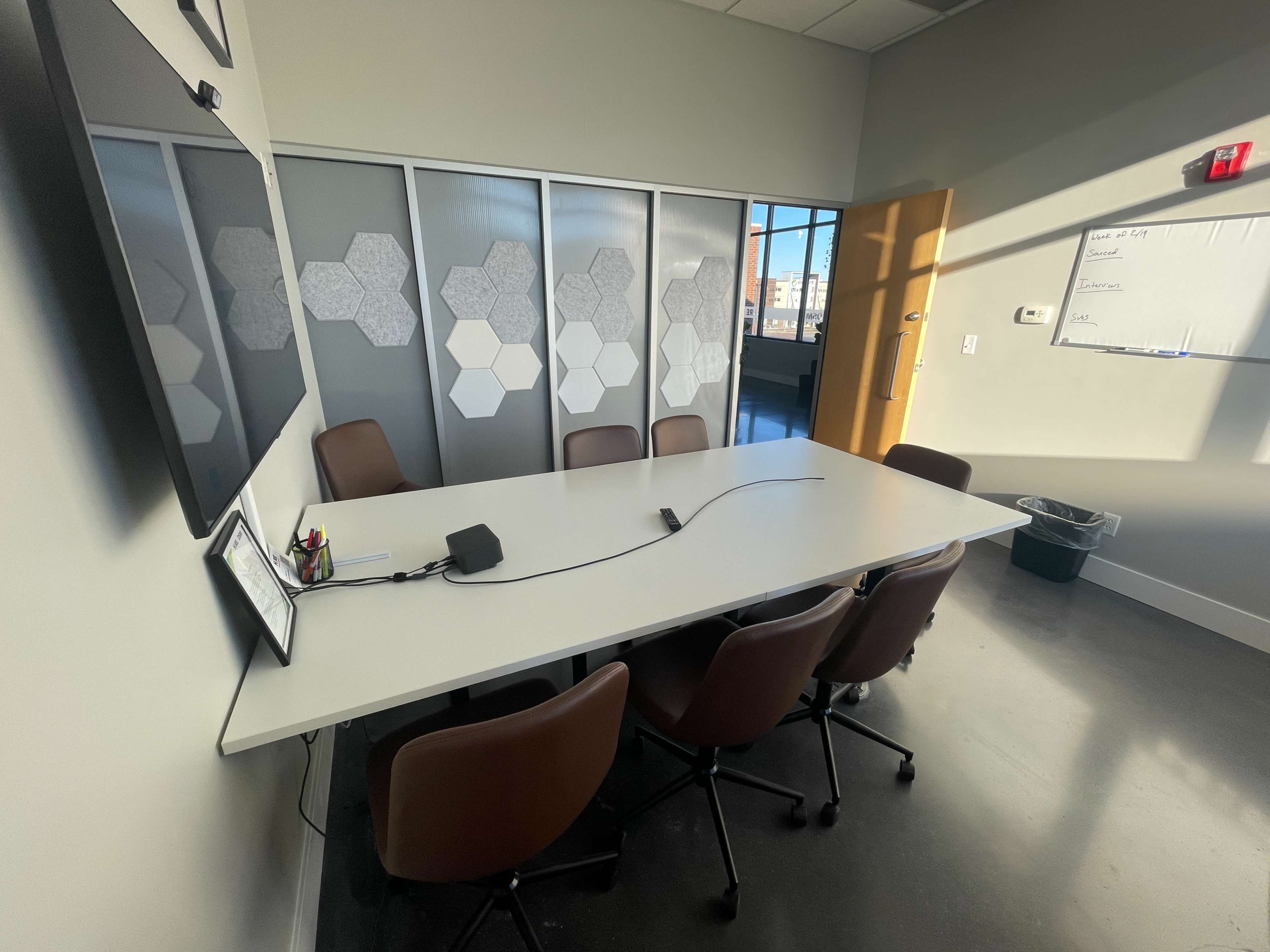 The image shows a modern conference room with a long table, several brown chairs, a wall-mounted screen, and a whiteboard.