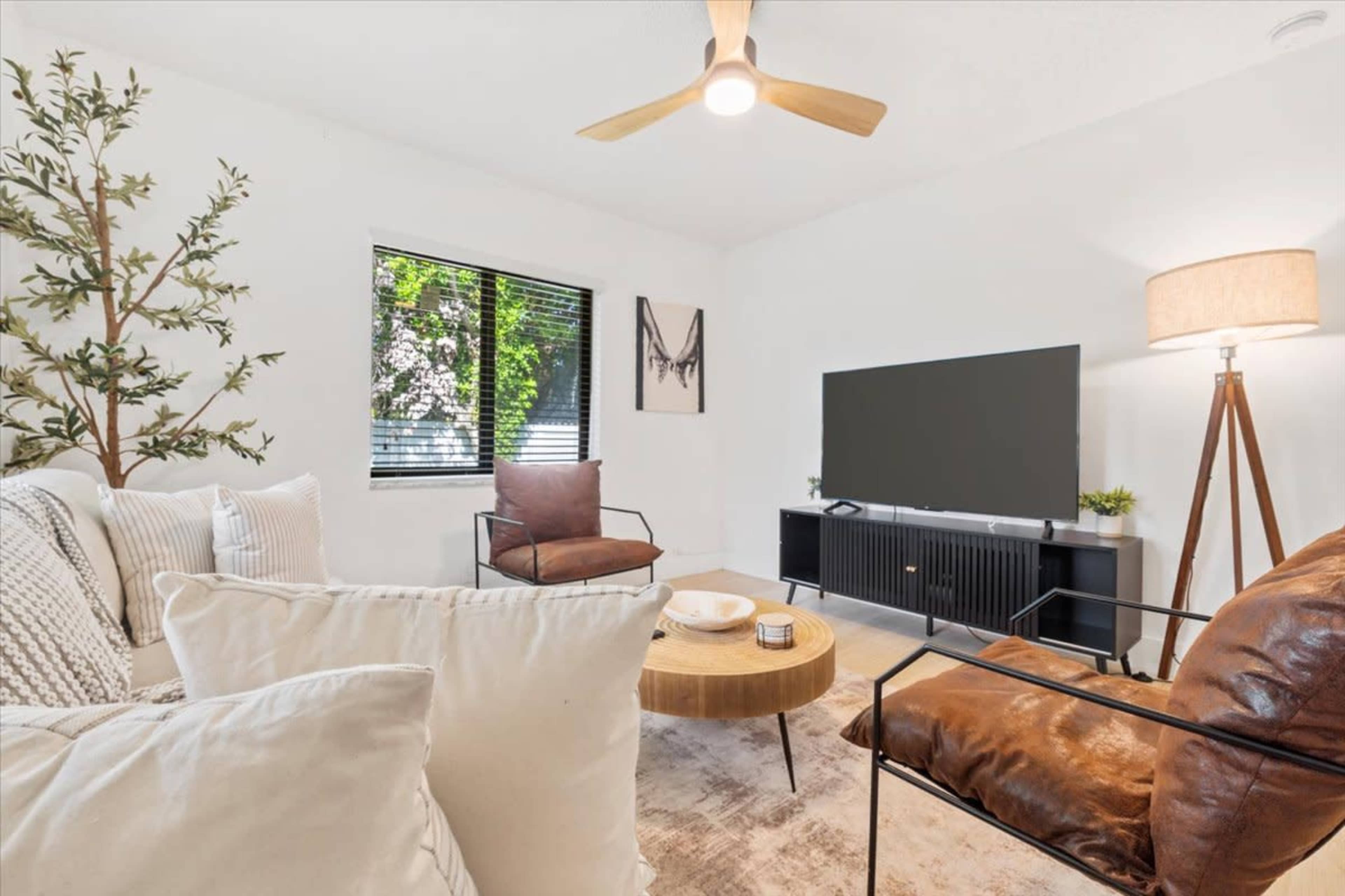 A well-lit living room features a beige couch, brown armchairs, a round wooden coffee table, and a large TV mounted on a black media console.