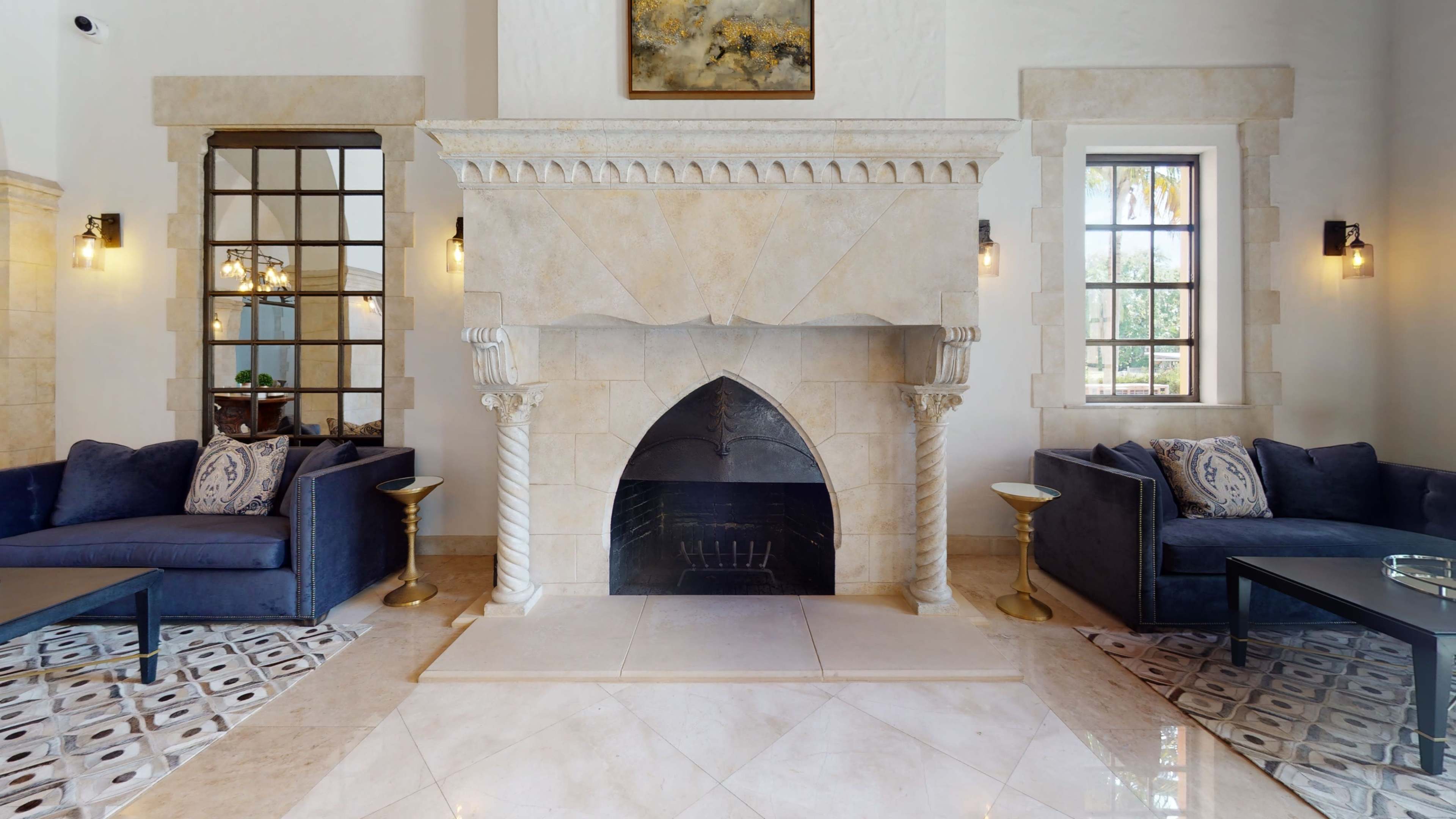 A spacious lobby with a stone fireplace at the center, flanked by two blue sofas and framed by large, decorative windows.