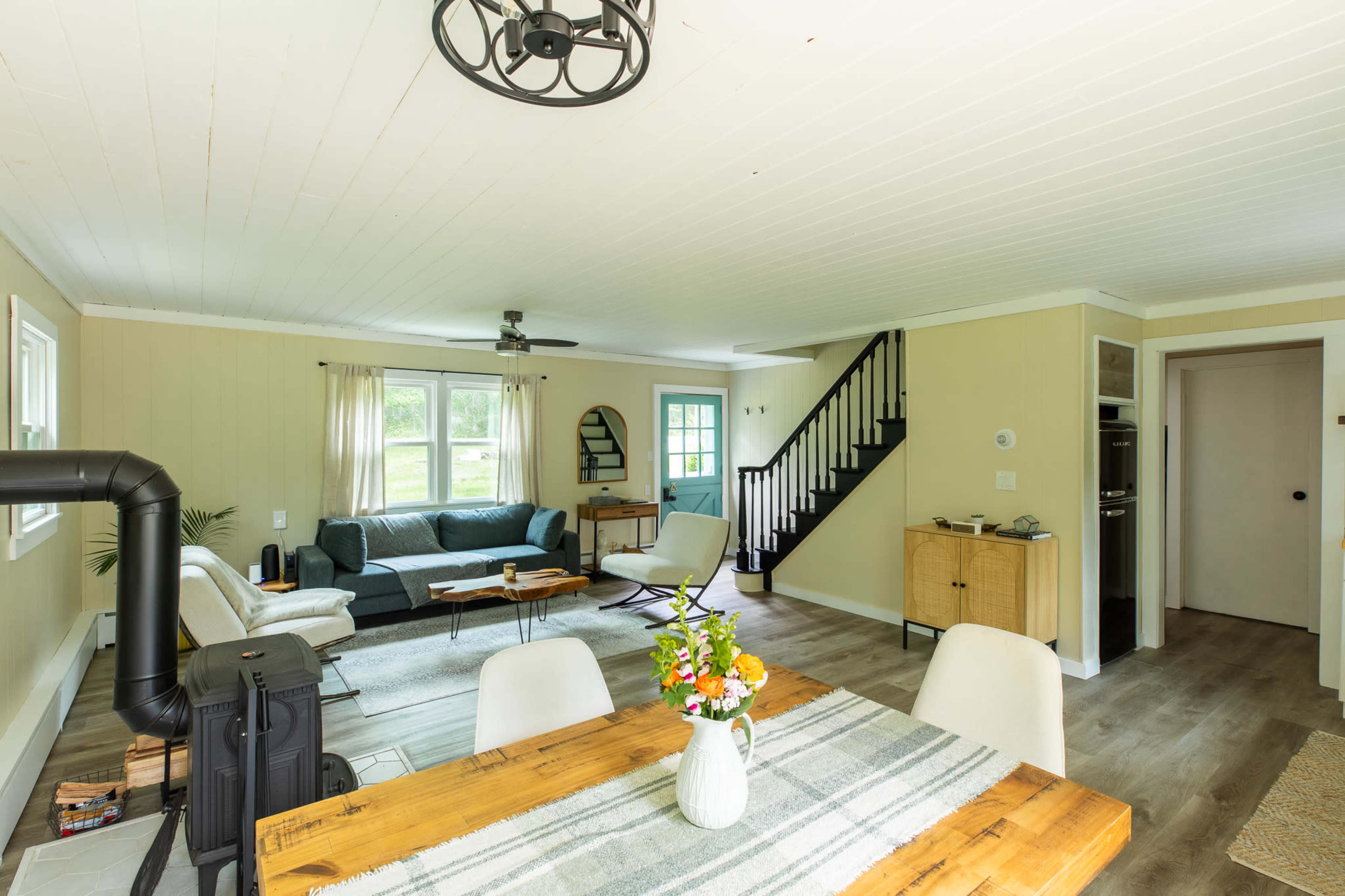 The image shows a light-filled living space that includes a seating area with a couch and armchair, a dining table, and a staircase leading to another level.