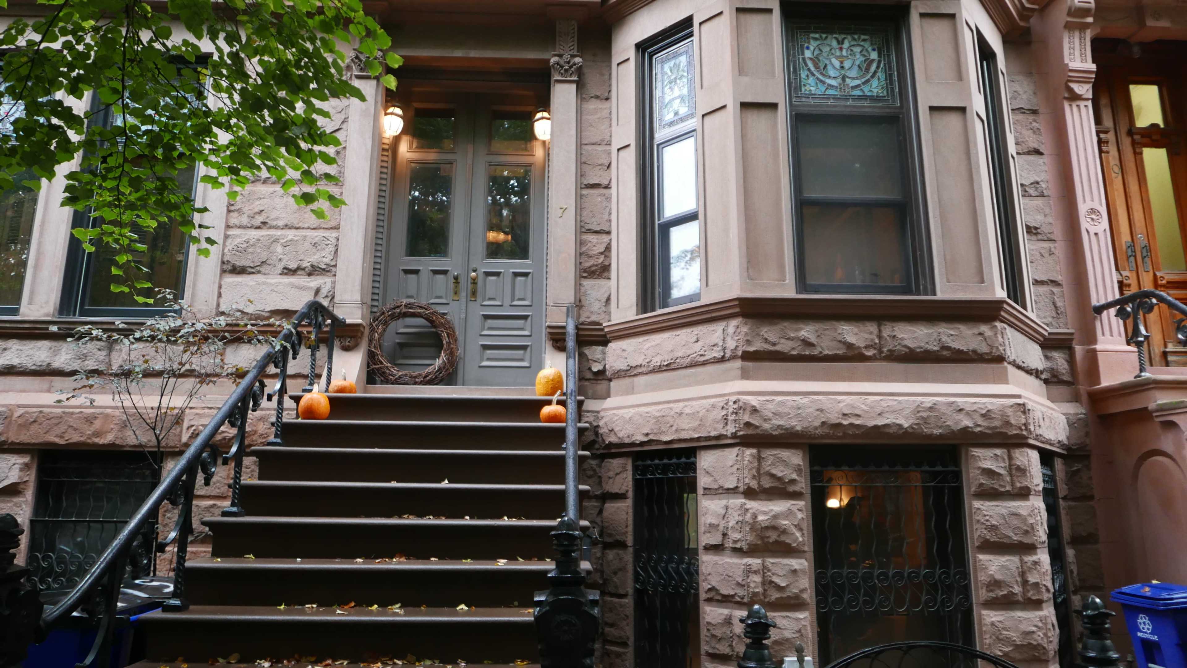 A set of steps leads to a brownstone entrance, adorned with a wreath and small pumpkins, surrounded by fall leaves and decorative elements.
