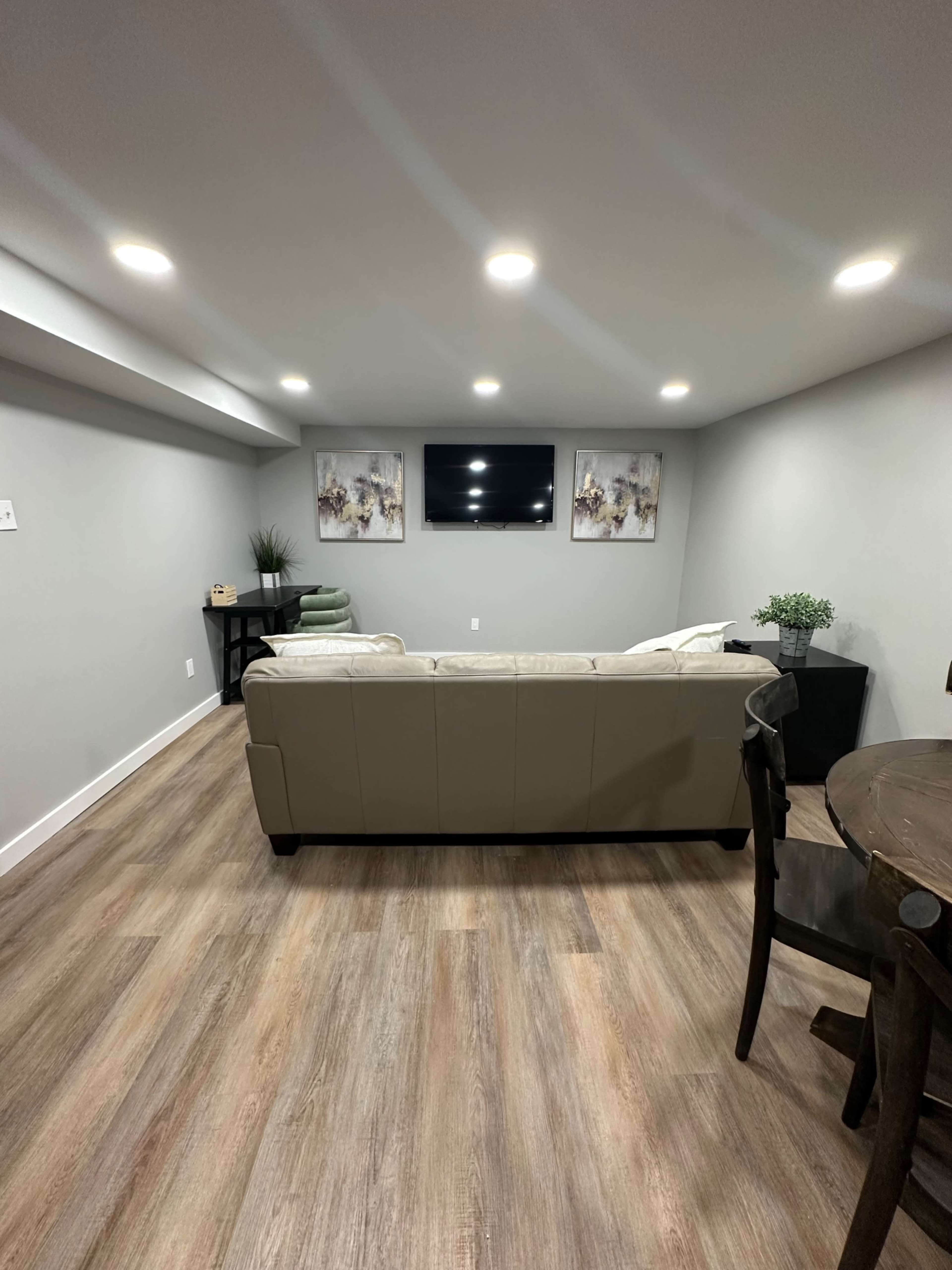 The image shows a modern basement living area featuring a light-colored couch facing a wall-mounted television, with artwork above and a dining table nearby.