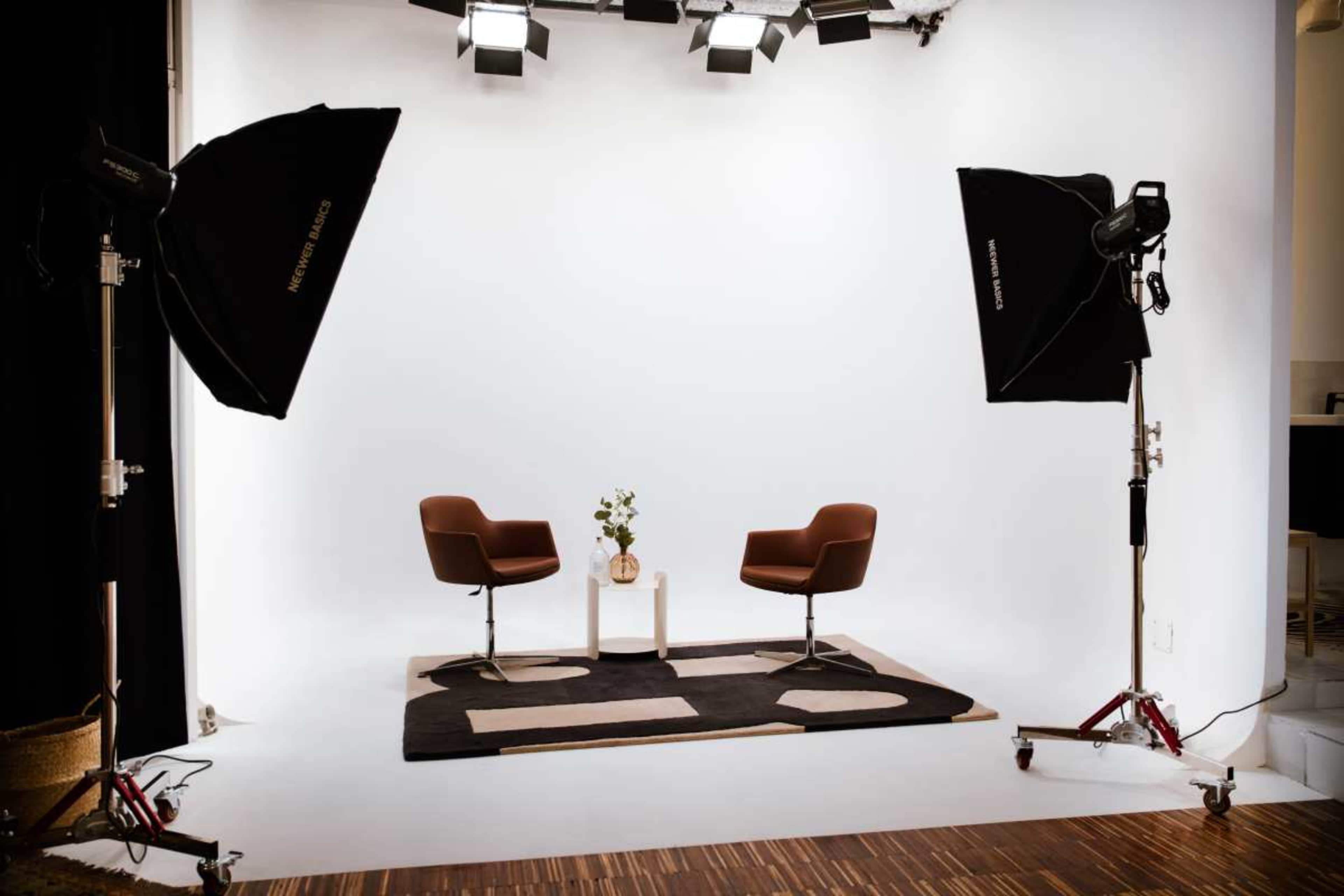 The image shows a minimalistic studio setup with two brown chairs facing each other on a patterned rug, flanked by large photography lights.