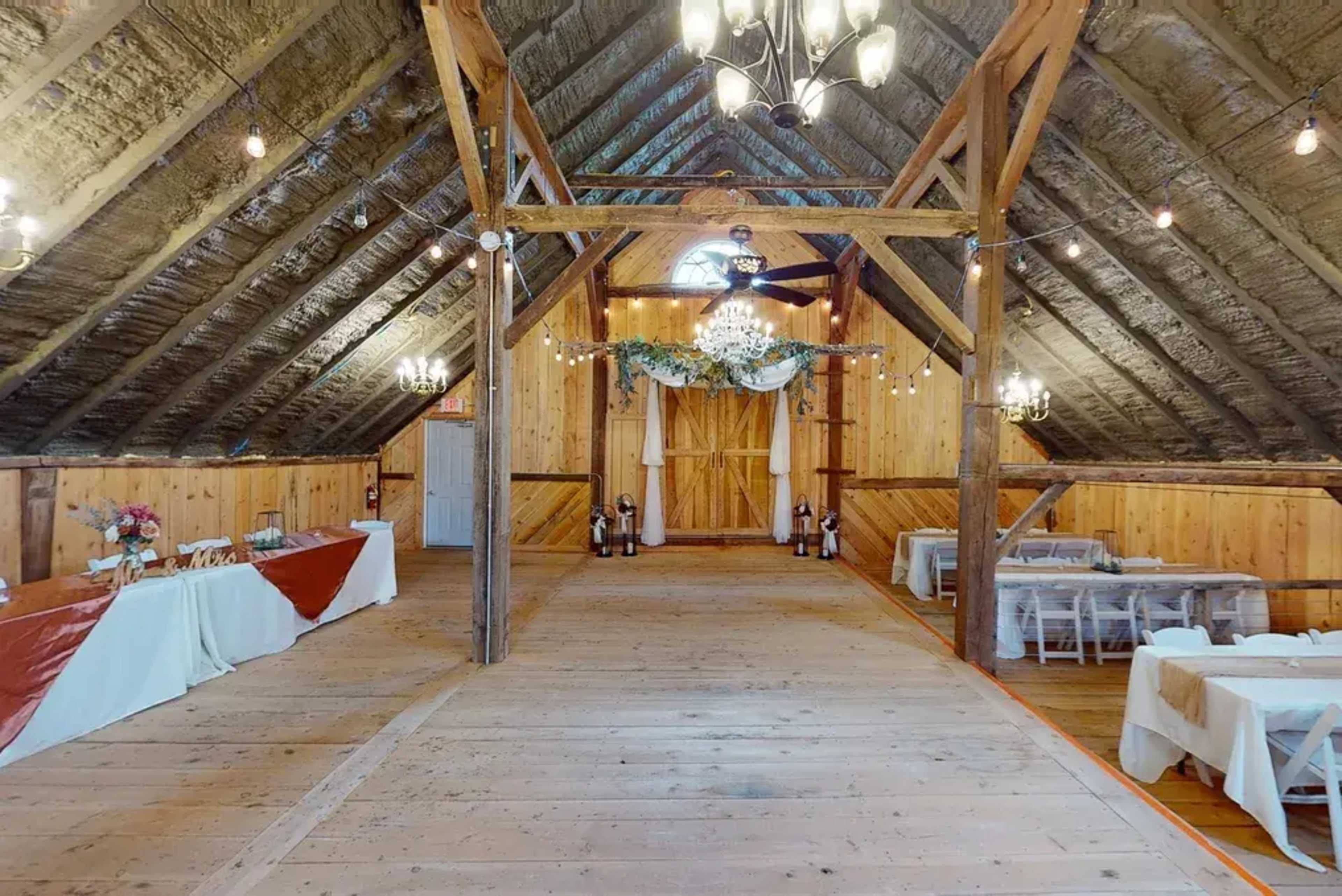 The interior of a rustic barn features wooden beams, a spacious central aisle, and tables set up for an event, with decorative lighting and floral arrangements.