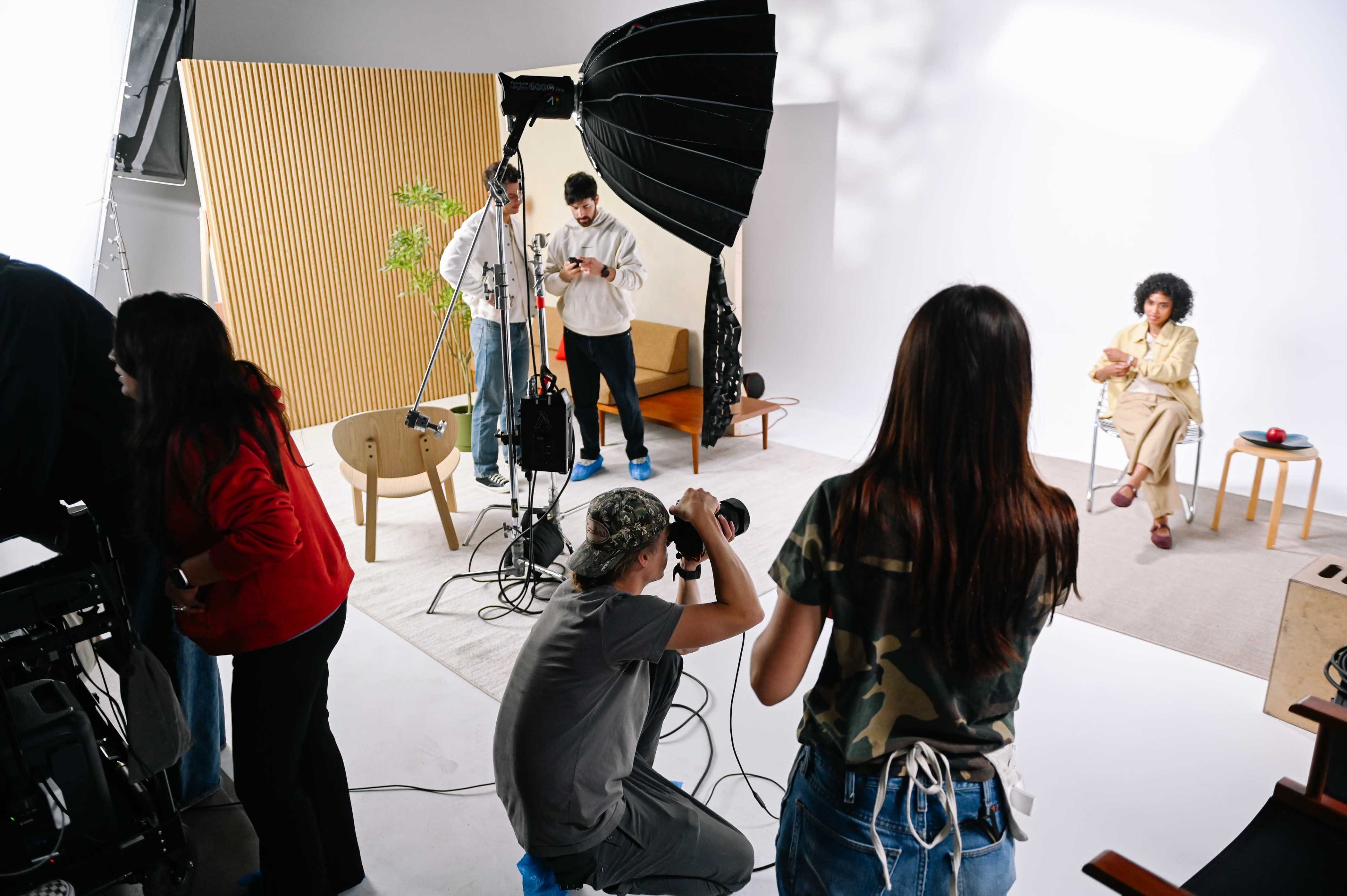 A television or photo shoot is taking place with a female subject seated in front of the camera while crew members adjust equipment and check their devices.