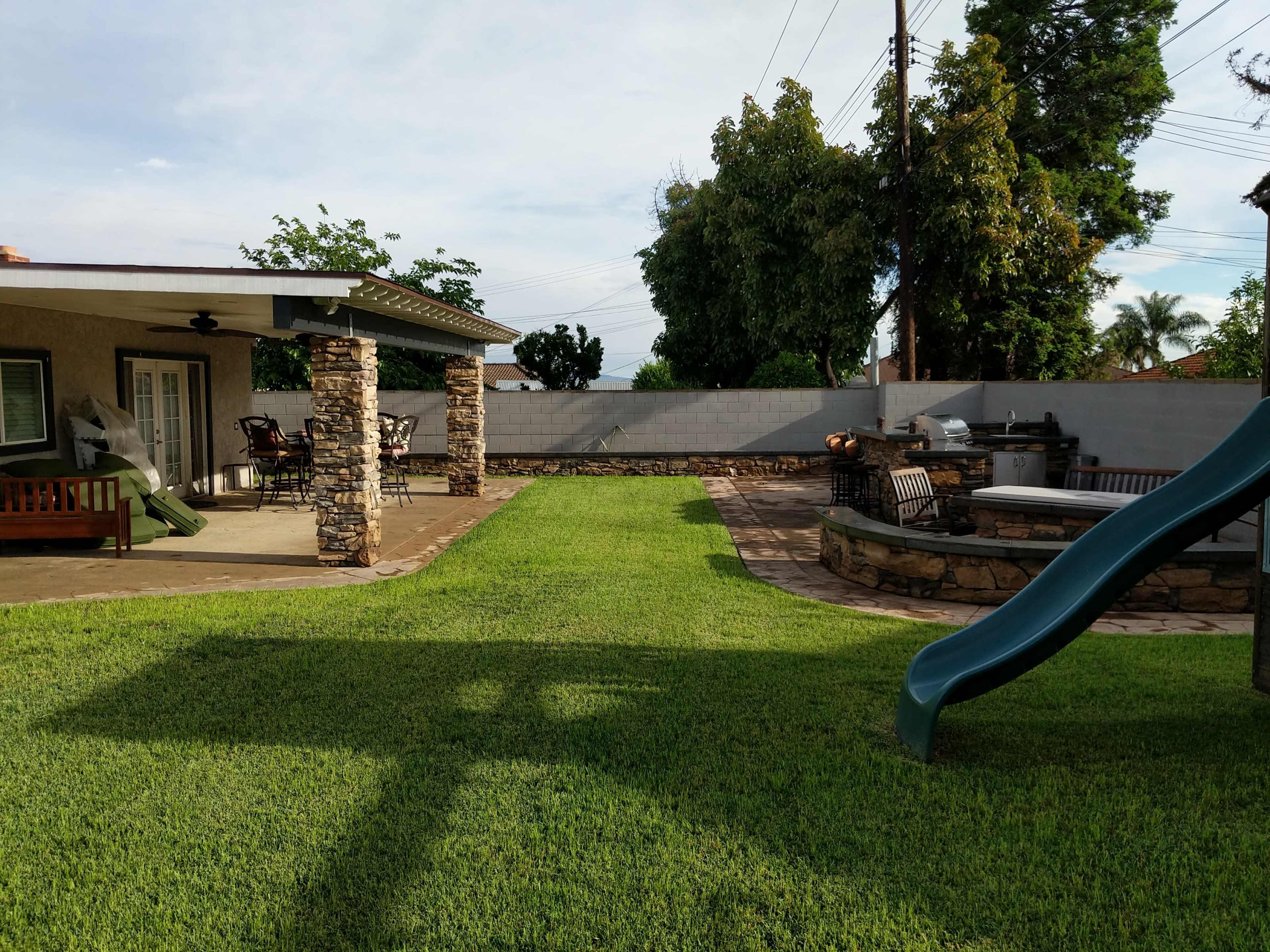 A backyard with a well-maintained green lawn, a patio area with stone pillars, and a slide next to a stone wall.