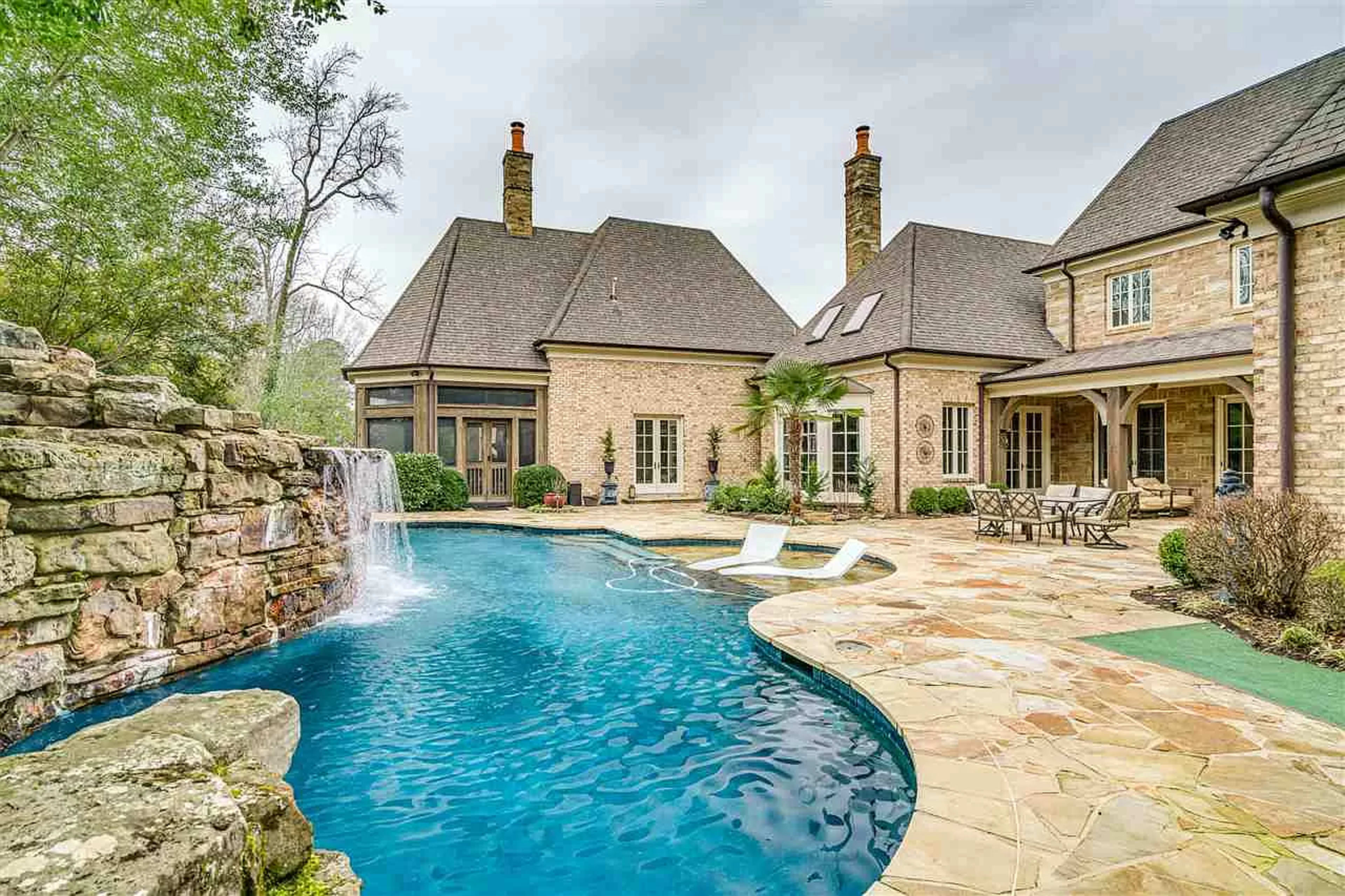 A backyard with a swimming pool, a stone waterfall, and a surrounding patio area with lounge chairs and seating arrangements.