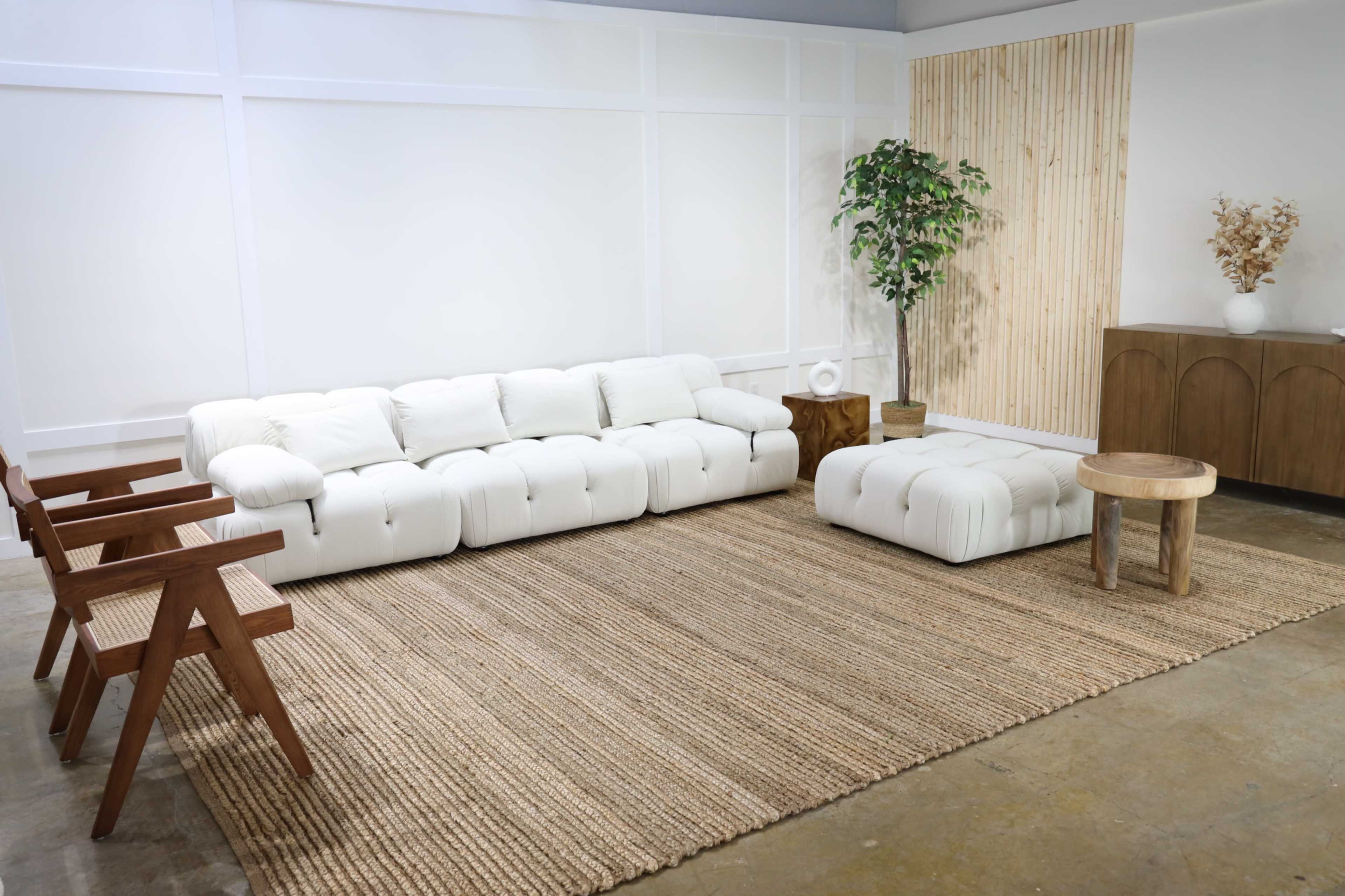 A spacious living area features a white sectional sofa, a matching ottoman, wooden chairs, and a round side table on a jute rug, with a potted plant and wooden wall accents in the background.