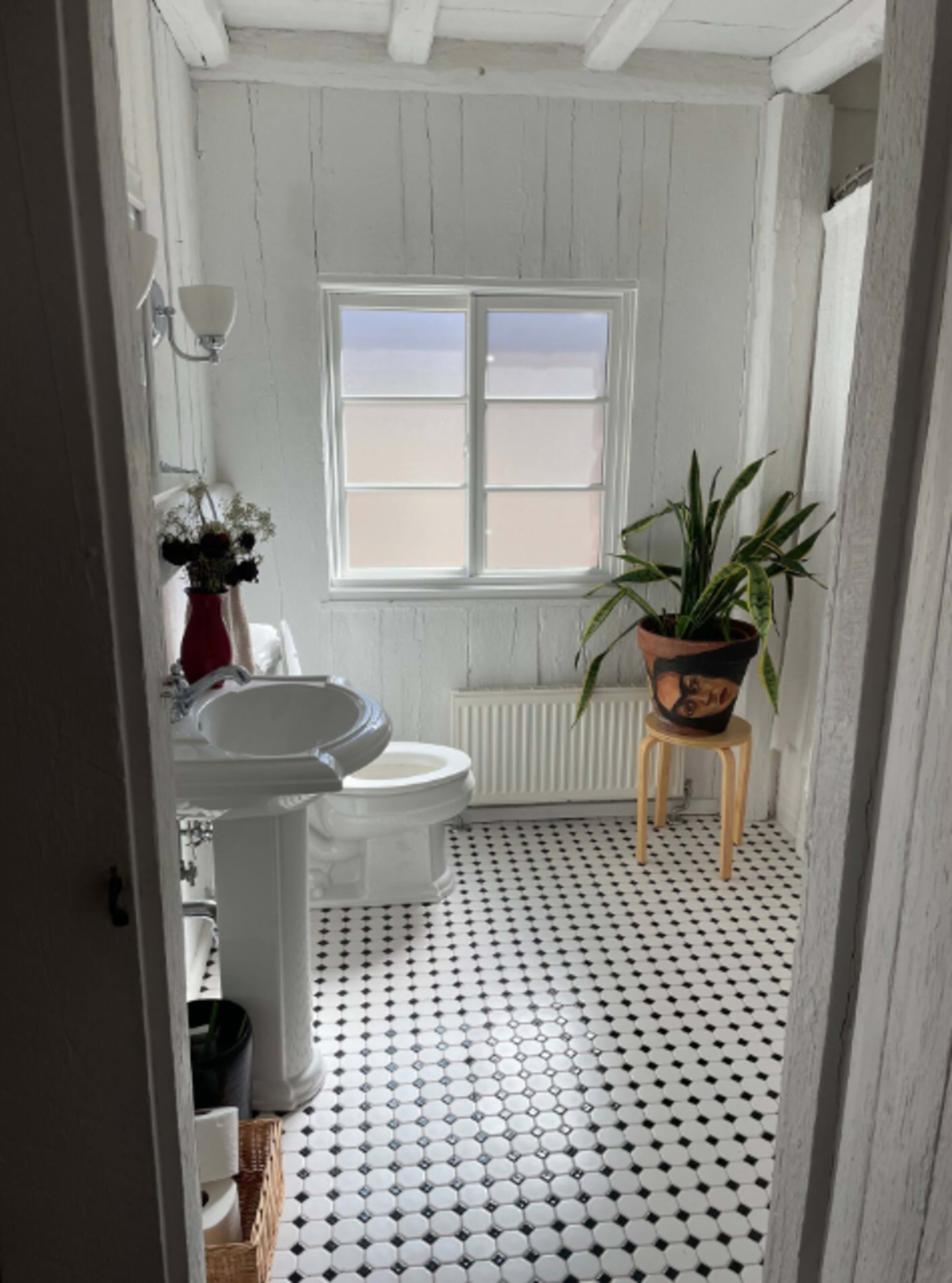 The image shows a bright bathroom with a white sink, a toilet, a potted plant on a stool, and patterned black-and-white tile flooring.