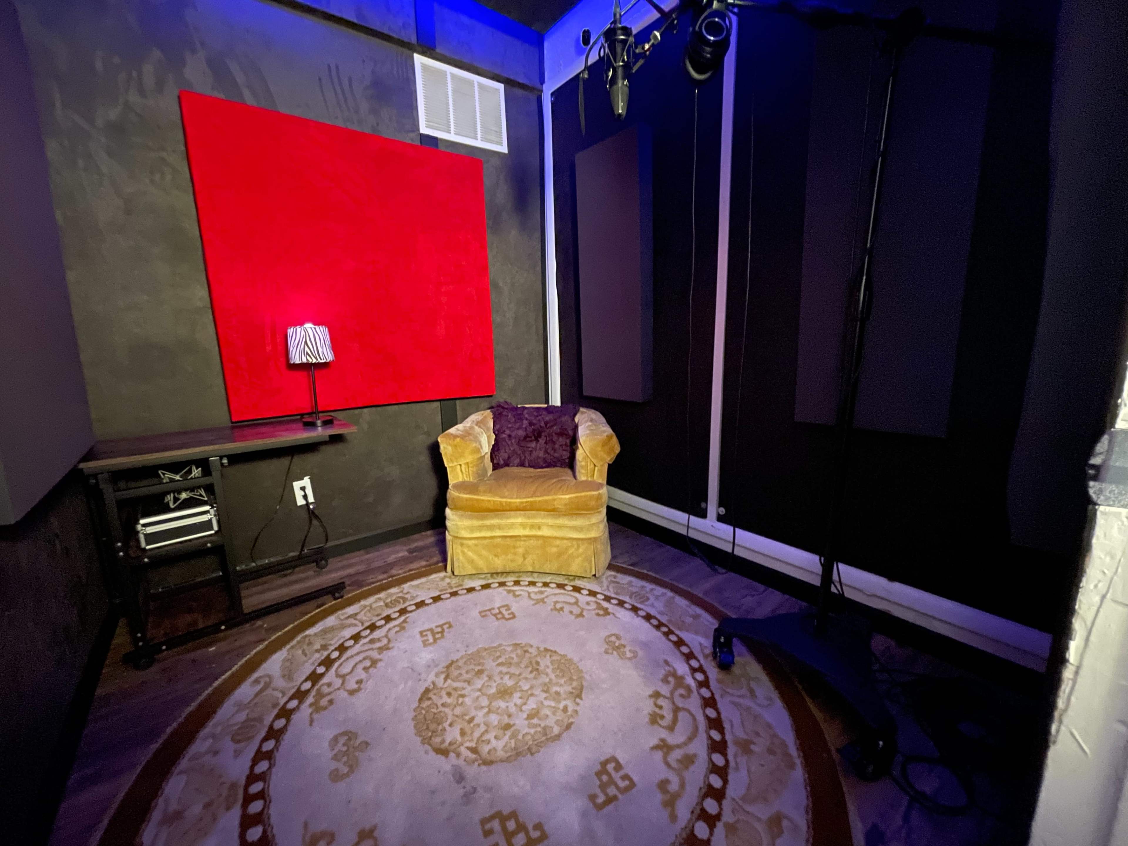 A small recording studio with a yellow armchair, a round carpet, and a red soundproof panel on the wall.