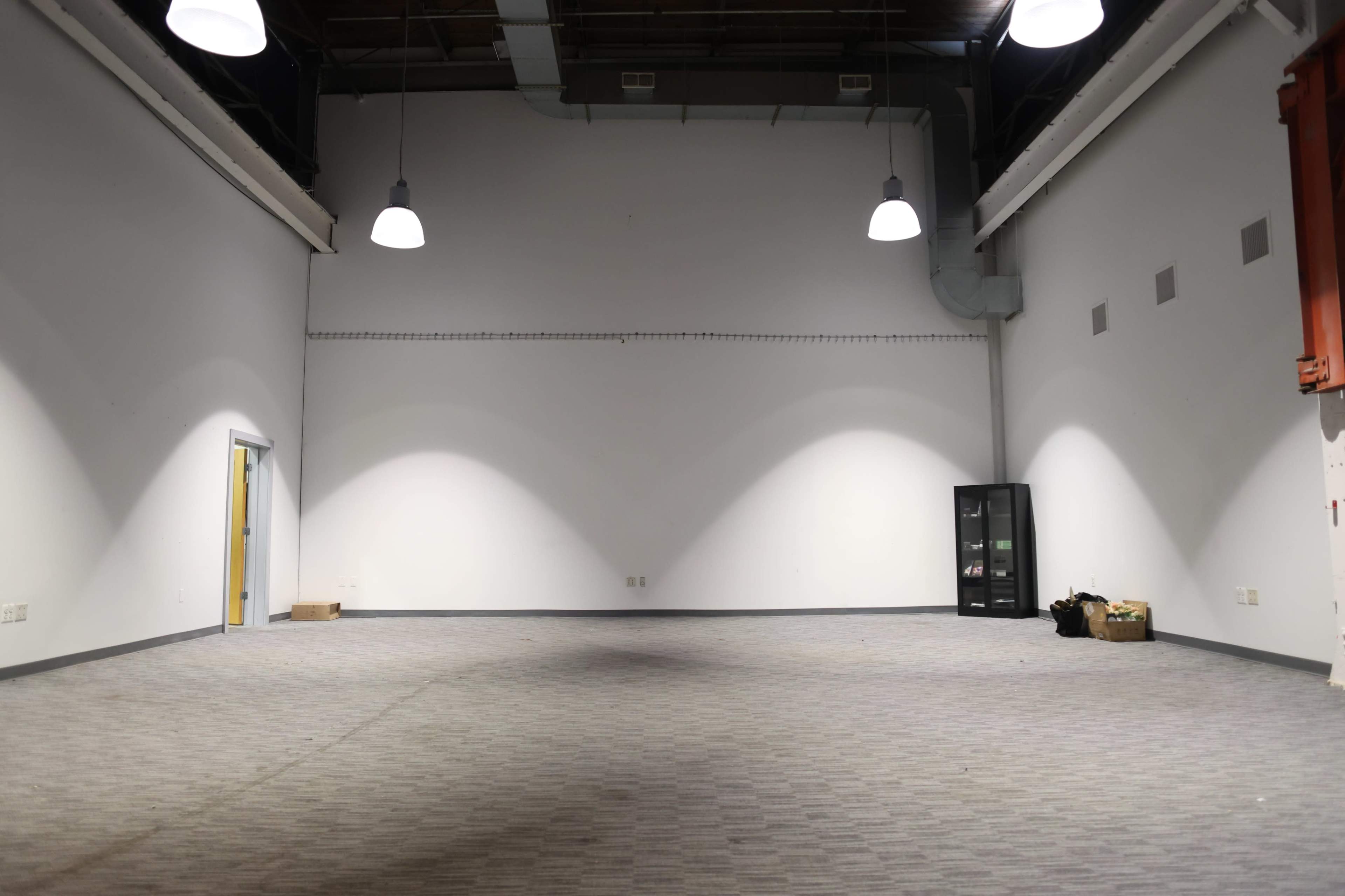 The image shows a large, empty room with gray carpet, minimal lighting from overhead lamps, and a few pieces of furniture against the walls.