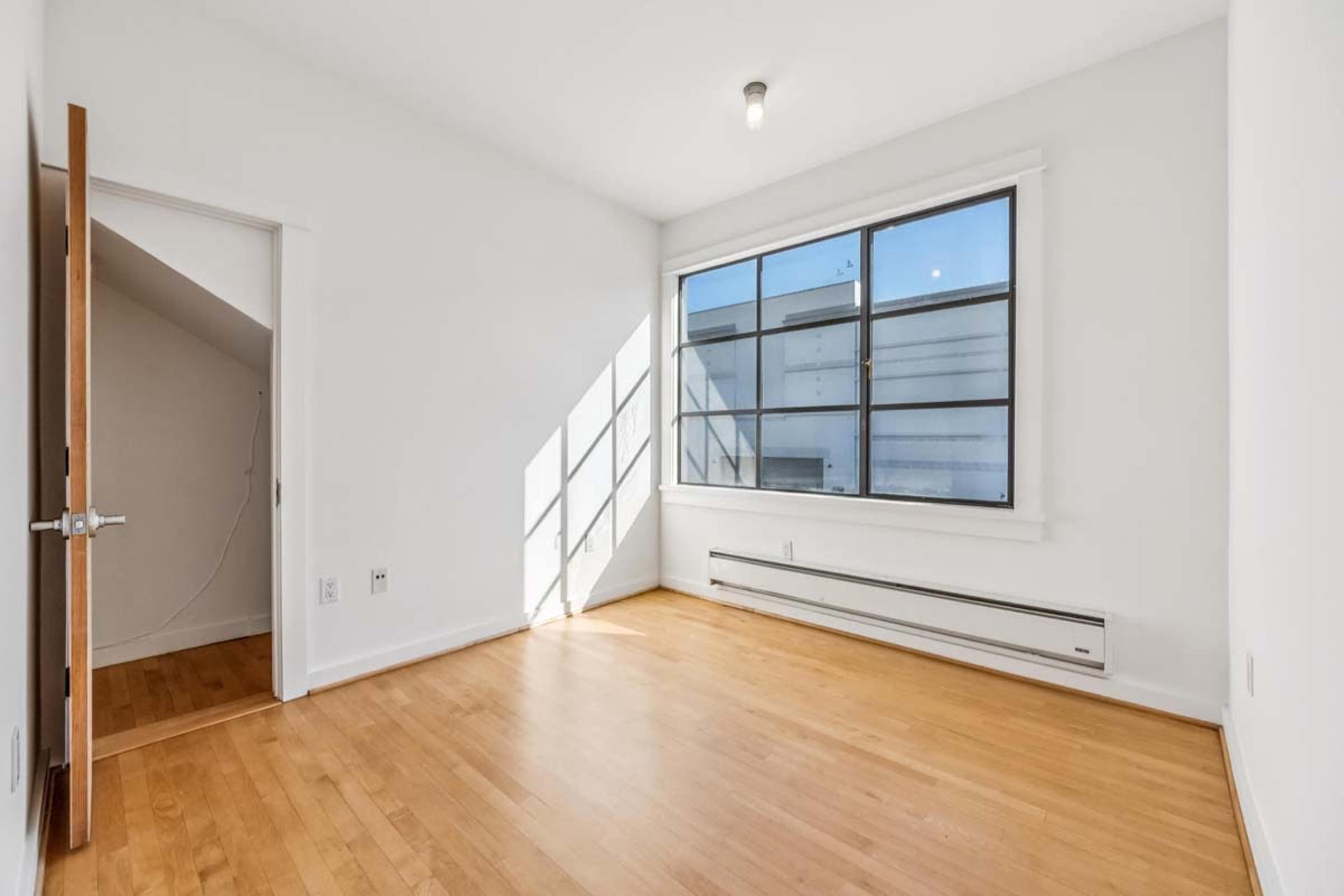 The image shows a vacant room with light wood flooring, a large window allowing natural light, and a simple white wall design.