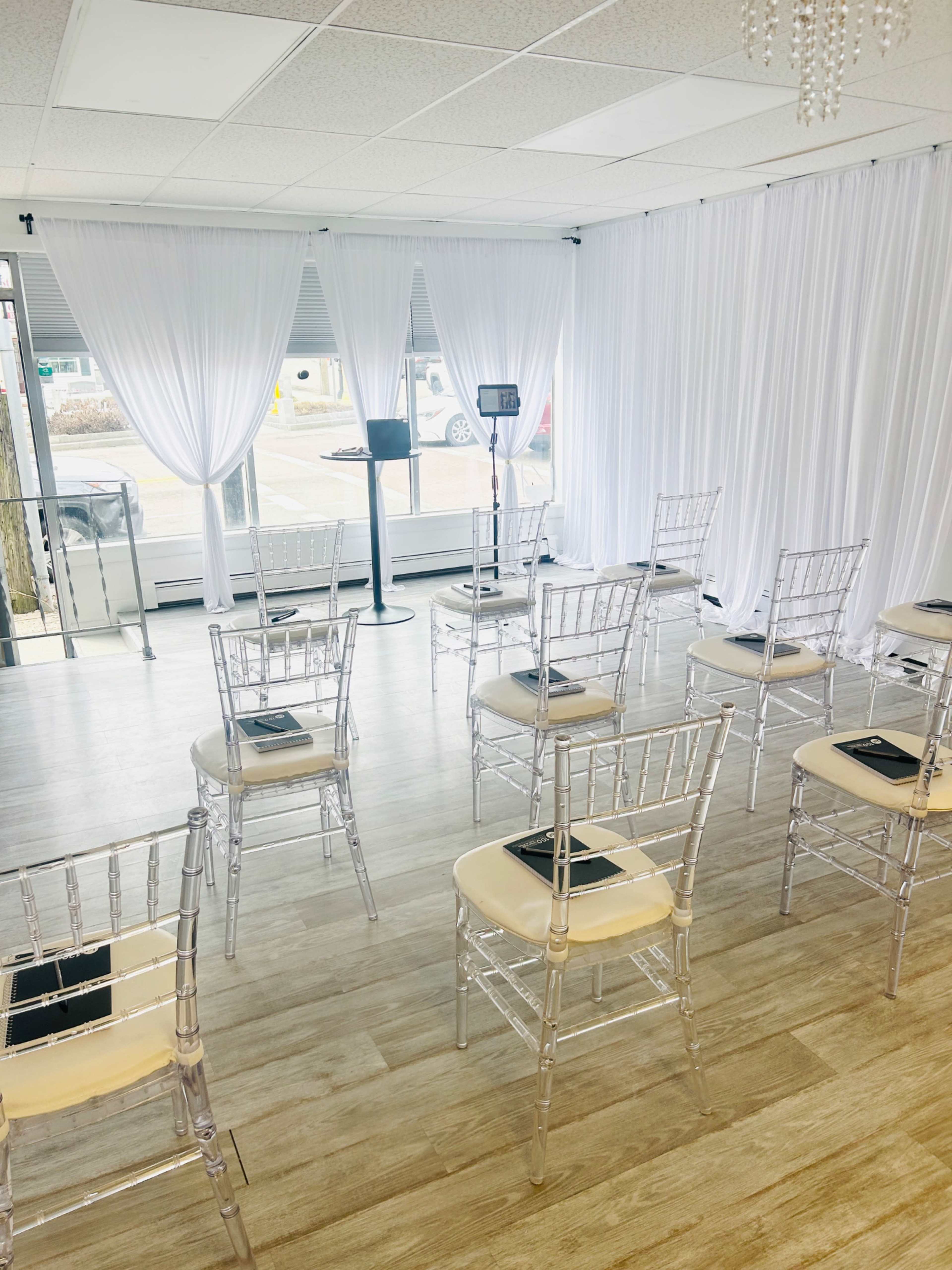 The image shows a spacious interior setting with transparent chairs arranged in rows, flanked by white draped curtains and natural light coming through large windows.