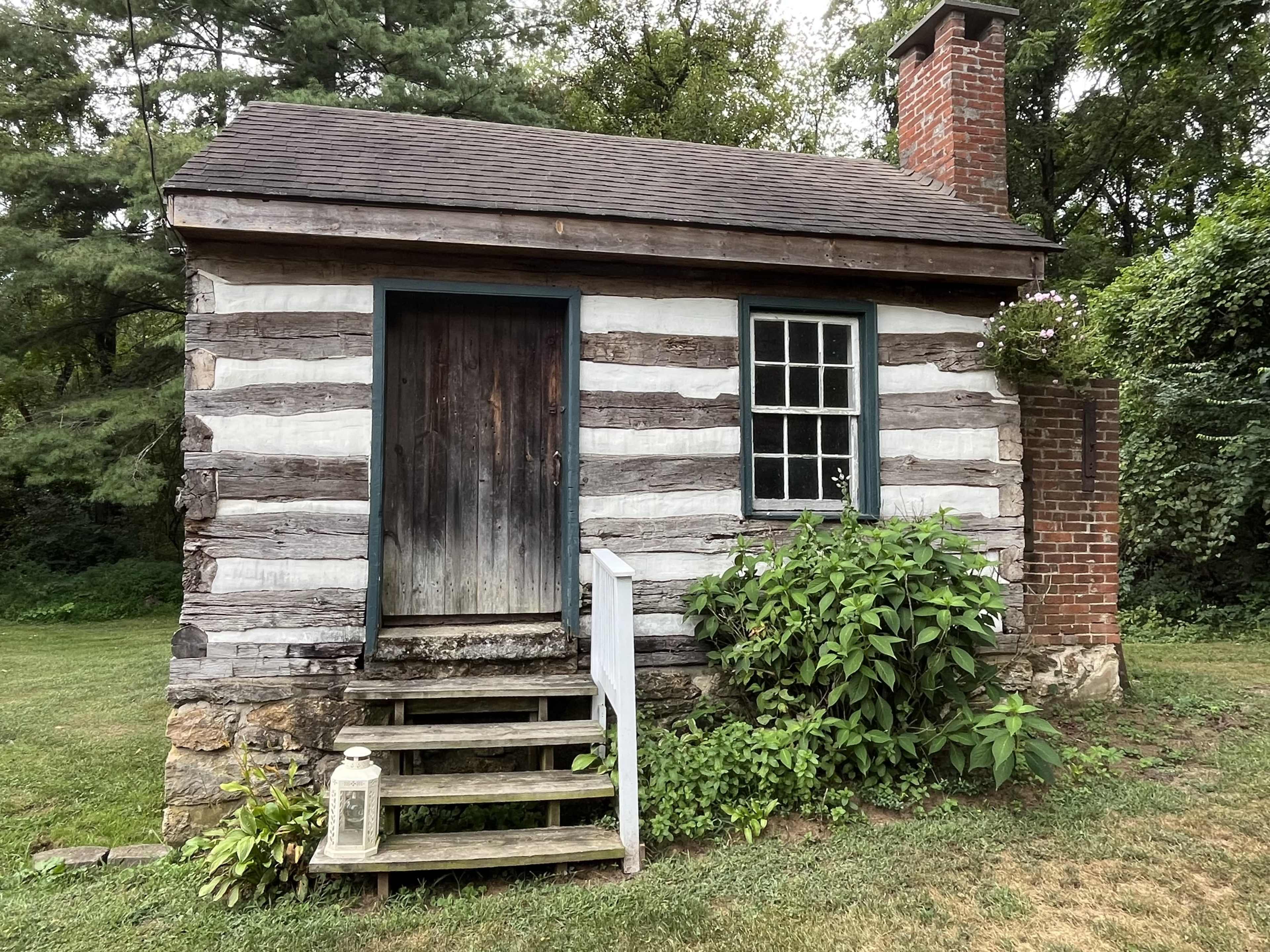 The image shows a small log cabin with a wooden front door, a front porch with steps, and a brick chimney, surrounded by greenery.