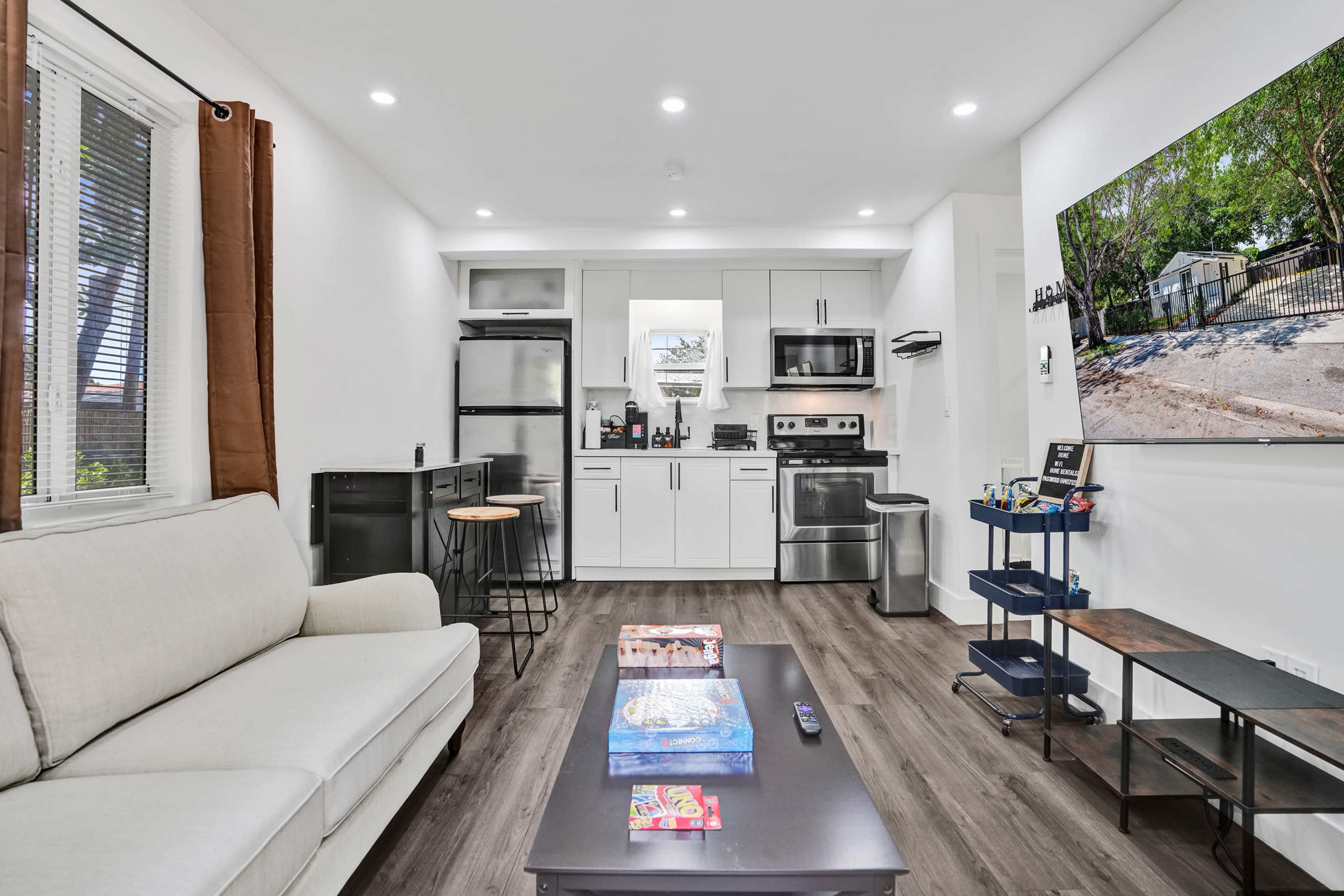 The image shows a modern living room with a beige sofa, a coffee table, a kitchenette with appliances, and a mounted television displaying an outdoor scene.
