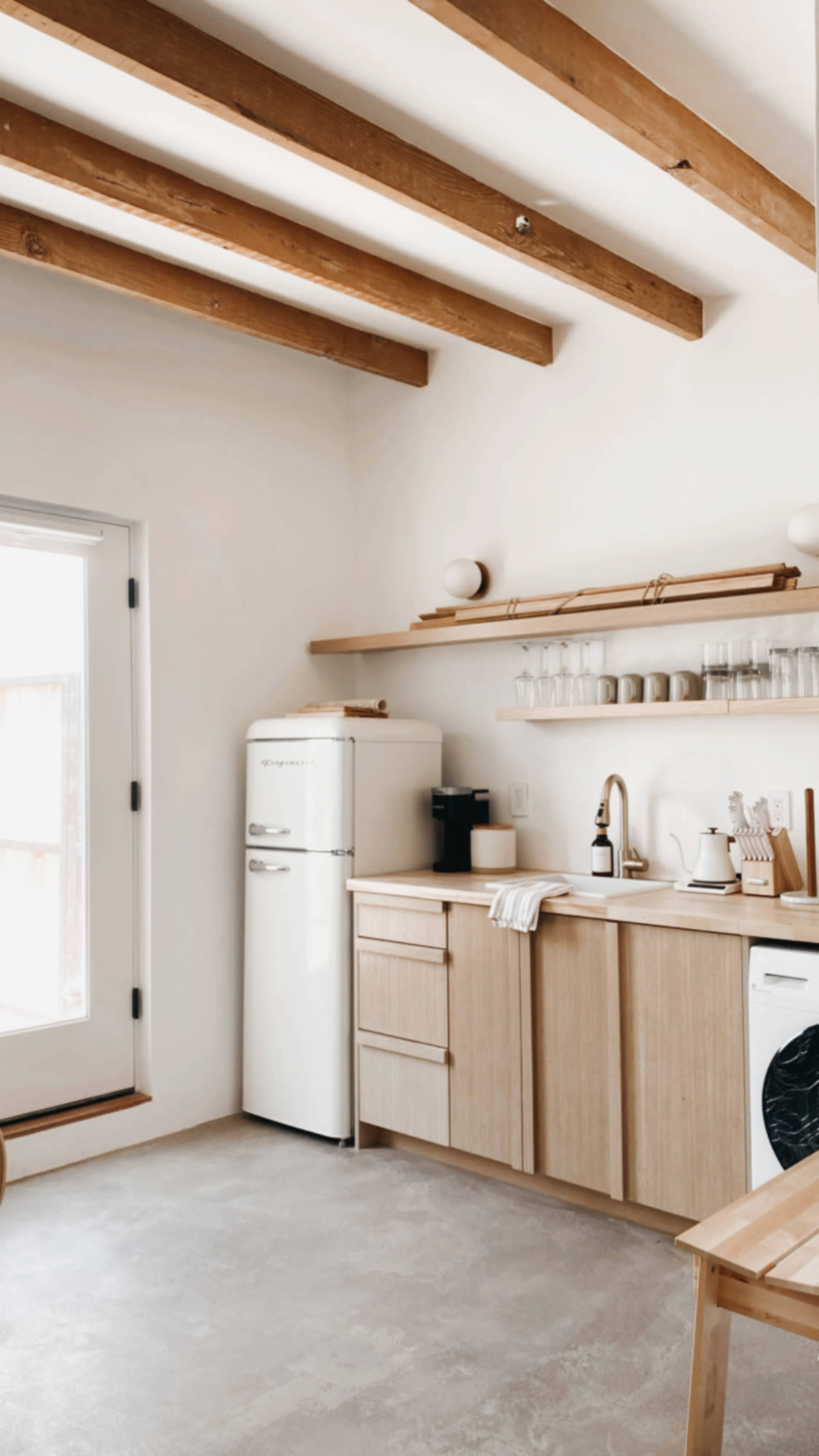 A modern kitchen features a white refrigerator, wooden cabinetry, and shelves with glassware and kitchen tools, illuminated by natural light from a nearby door.