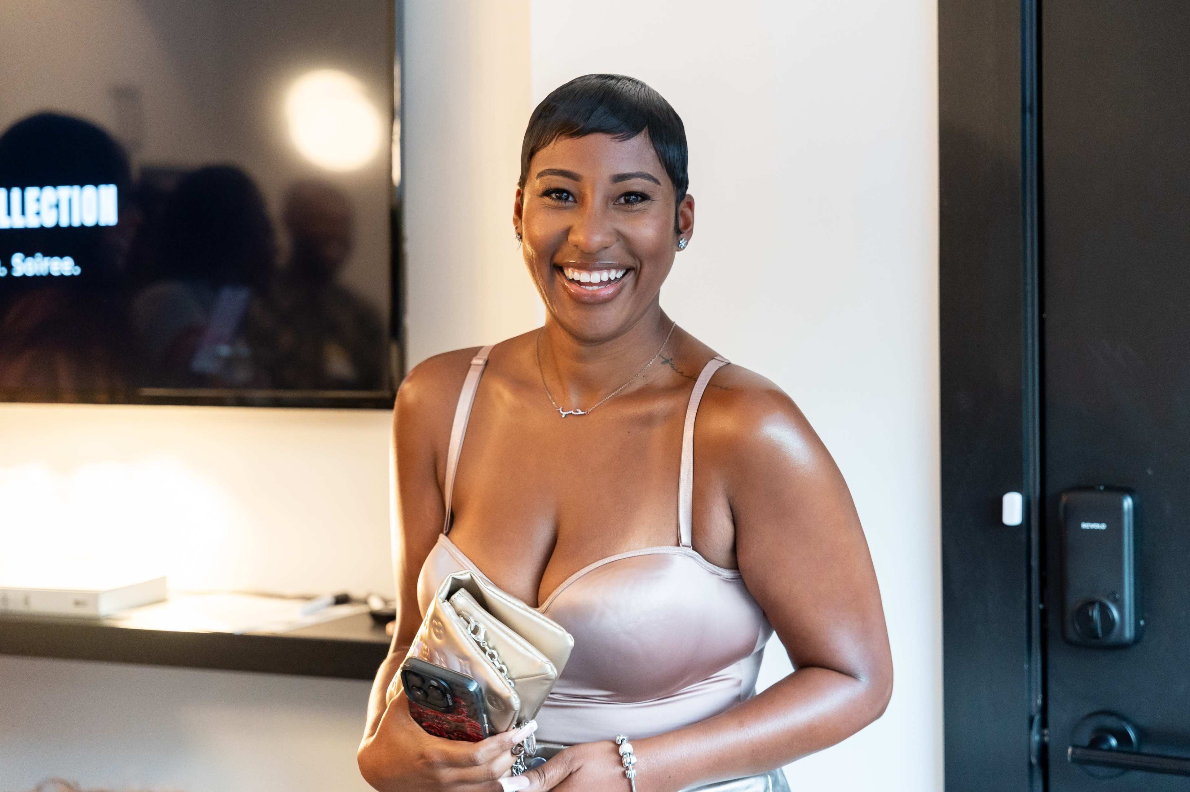 A woman with short hair and a light-colored dress smiles while holding a clutch bag in a modern indoor setting.