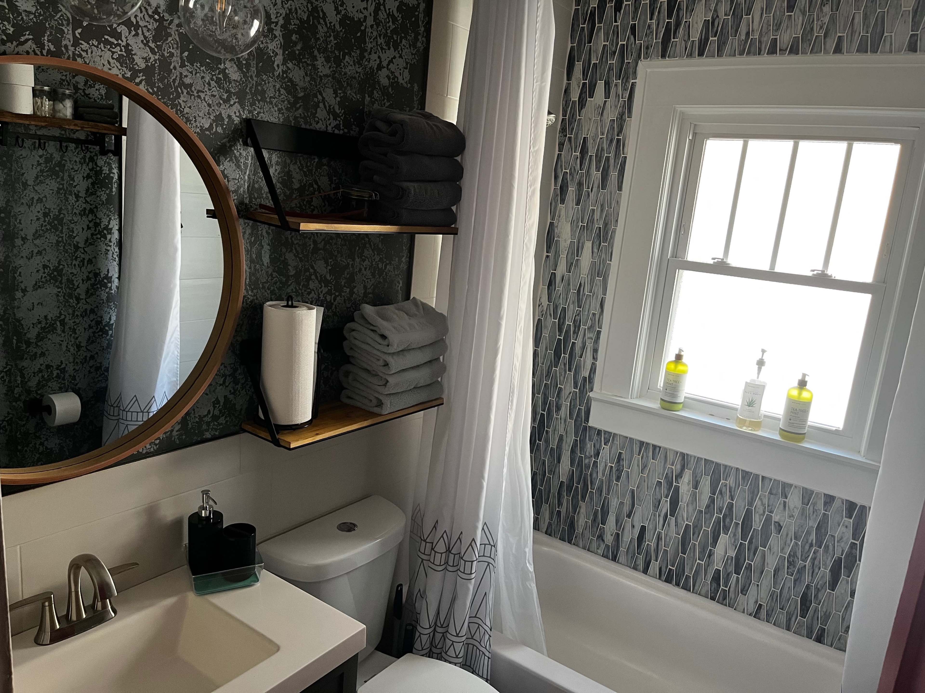 A modern bathroom featuring a round mirror, a bathtub, and shelves with neatly stacked towels and toiletries beside a window.