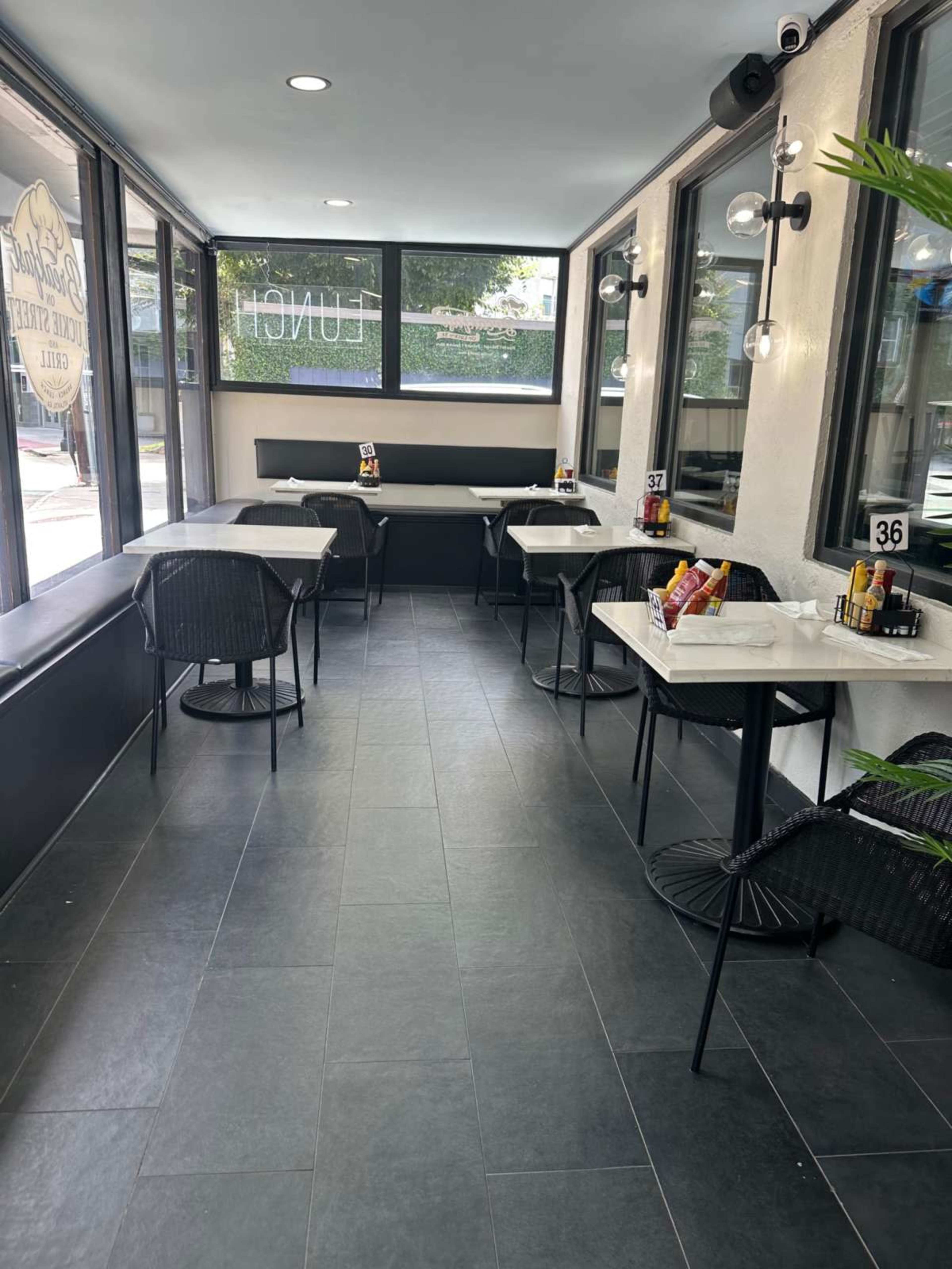 The image shows a modern, bright dining area with four tables, each set with condiments, in a sunlit space surrounded by large windows.