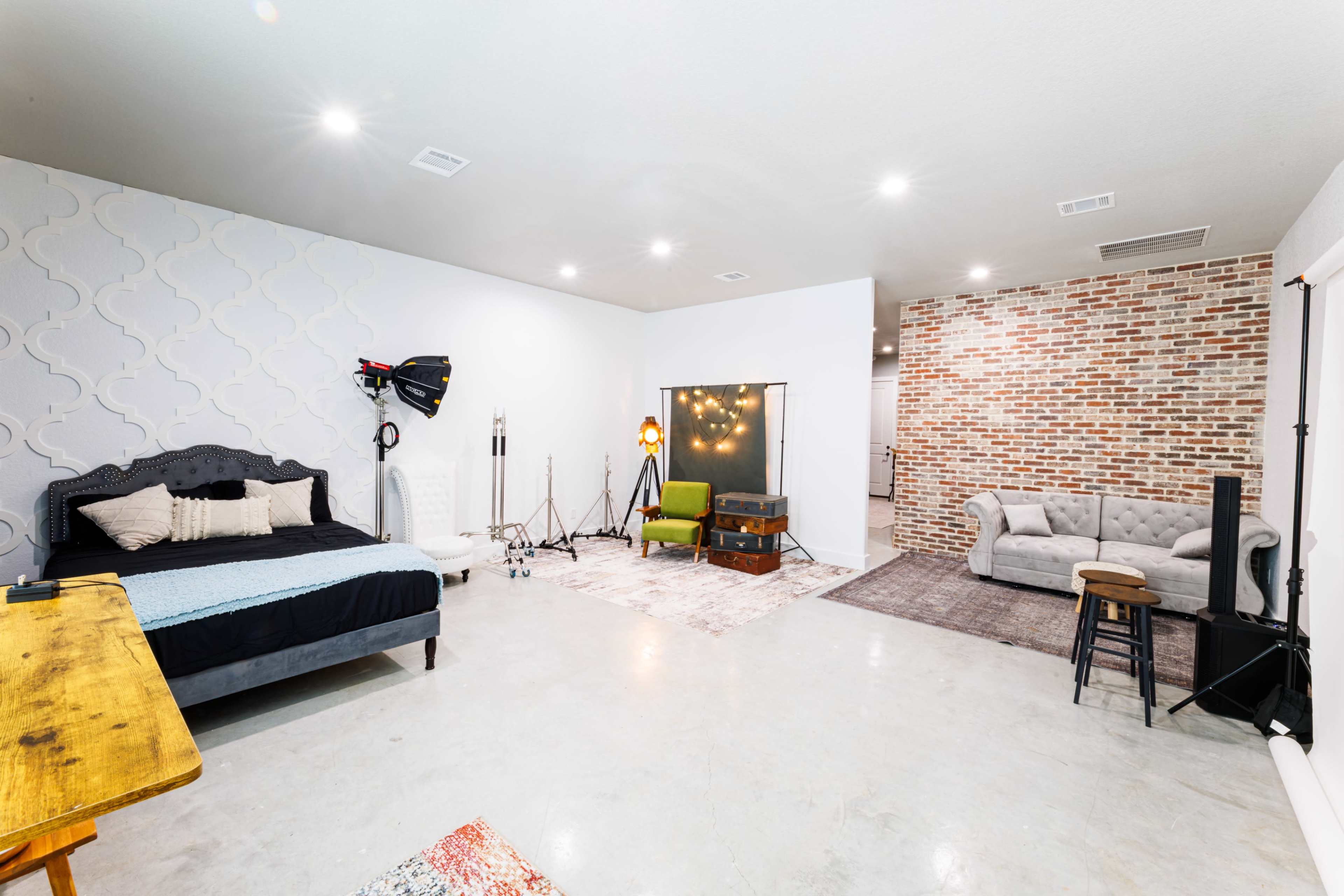 The image shows a spacious, modern room featuring a bed, a couch, various lighting equipment, and exposed brick walls.