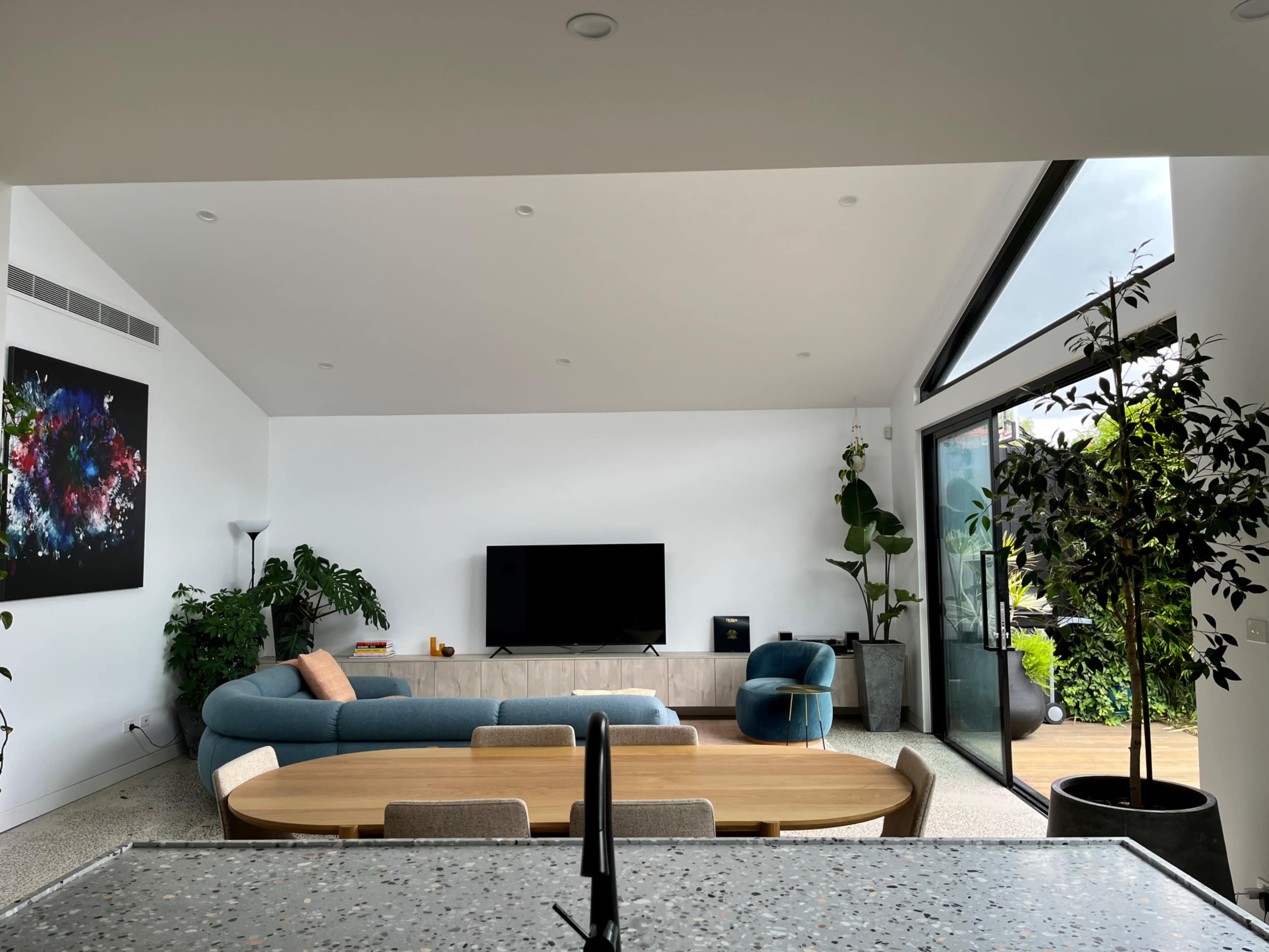 A modern living room with a sectional sofa, a large TV mounted on the wall, and plants near the windows.