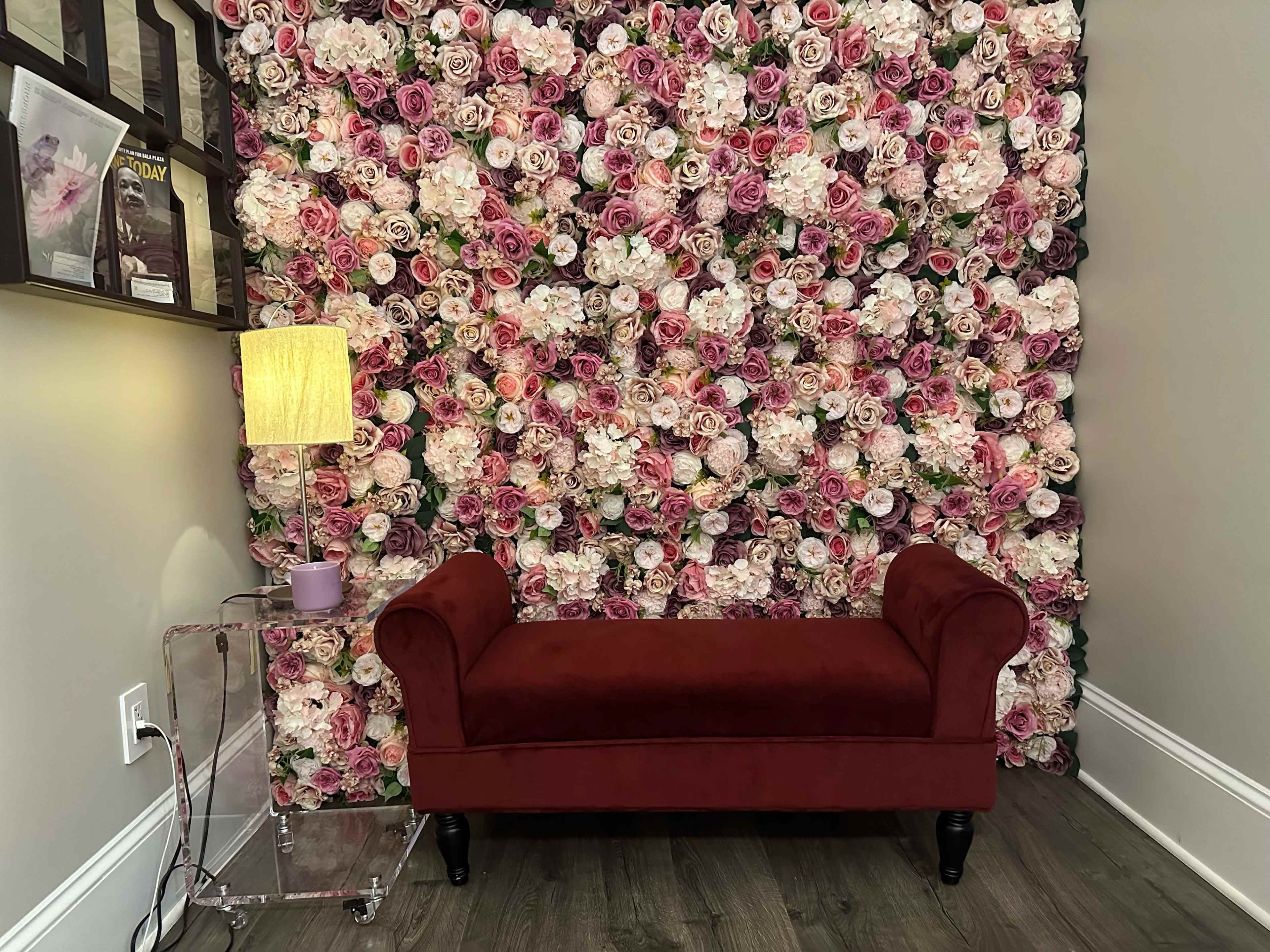 The image shows a velvet red bench in front of a wall covered with a dense arrangement of pink and white artificial flowers.
