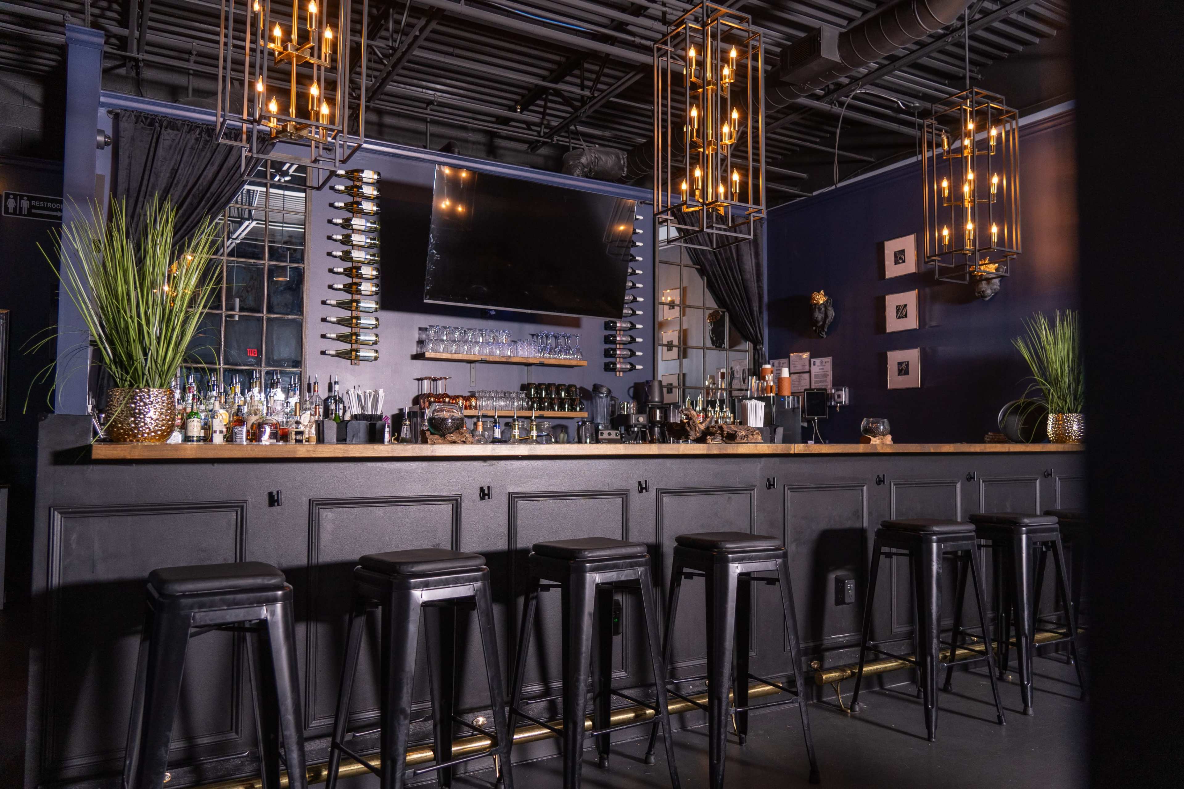 A modern bar area with black stools, a wooden counter, and decorative lighting fixtures, set against dark walls and a large screen.