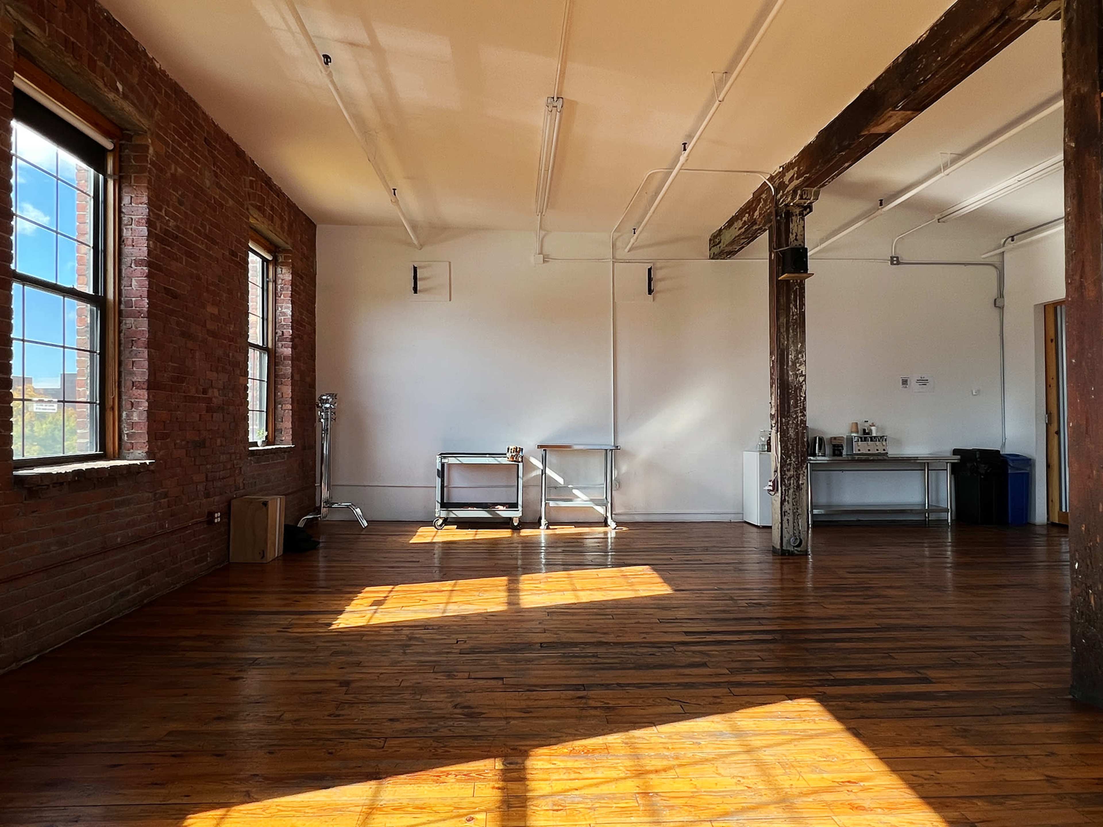 A spacious, sunlit room with wooden floors, exposed brick walls, and a few pieces of furniture including tables and appliances.
