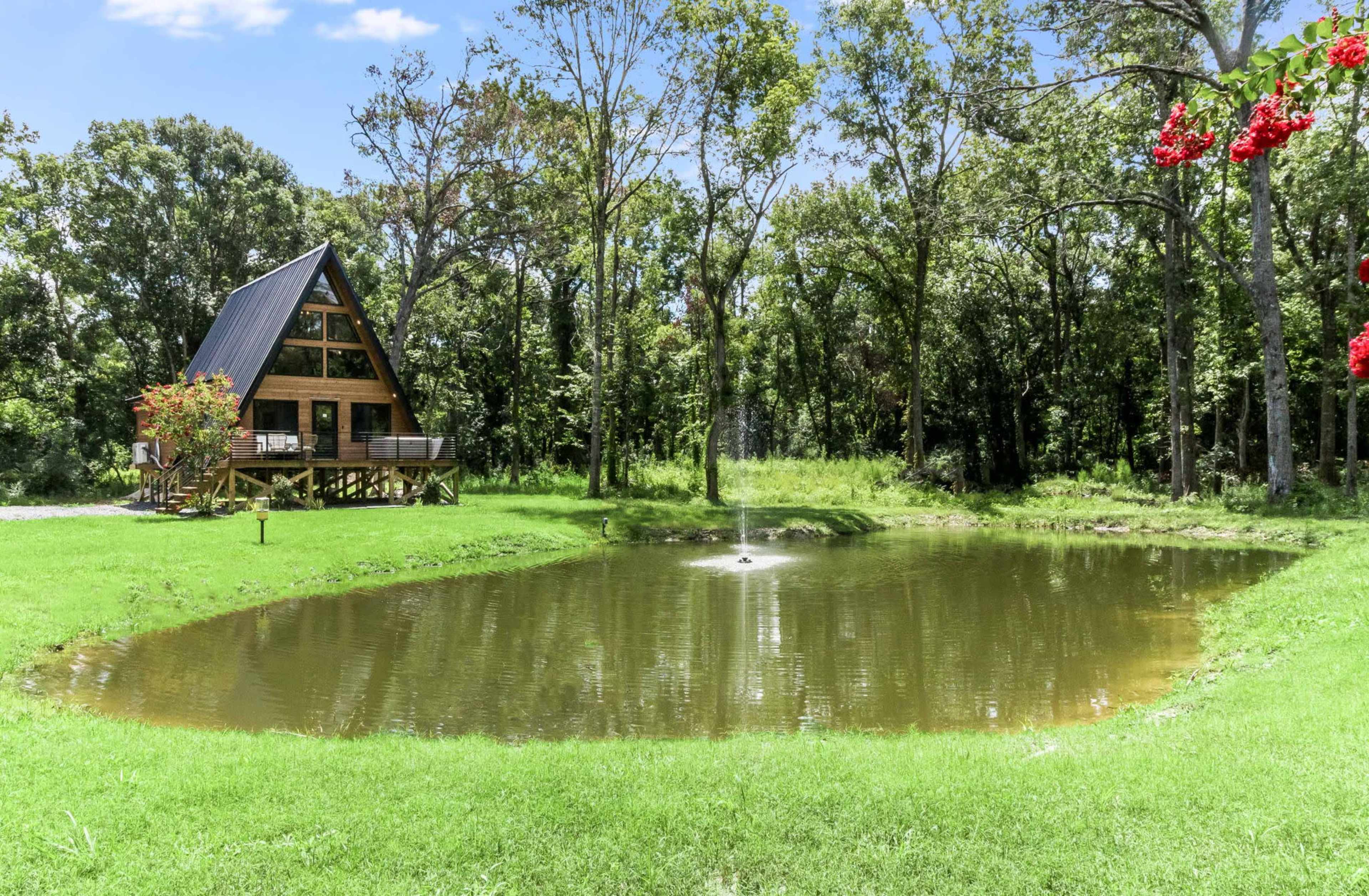 A wooden A-frame cabin beside a pond surrounded by trees and grassy land.