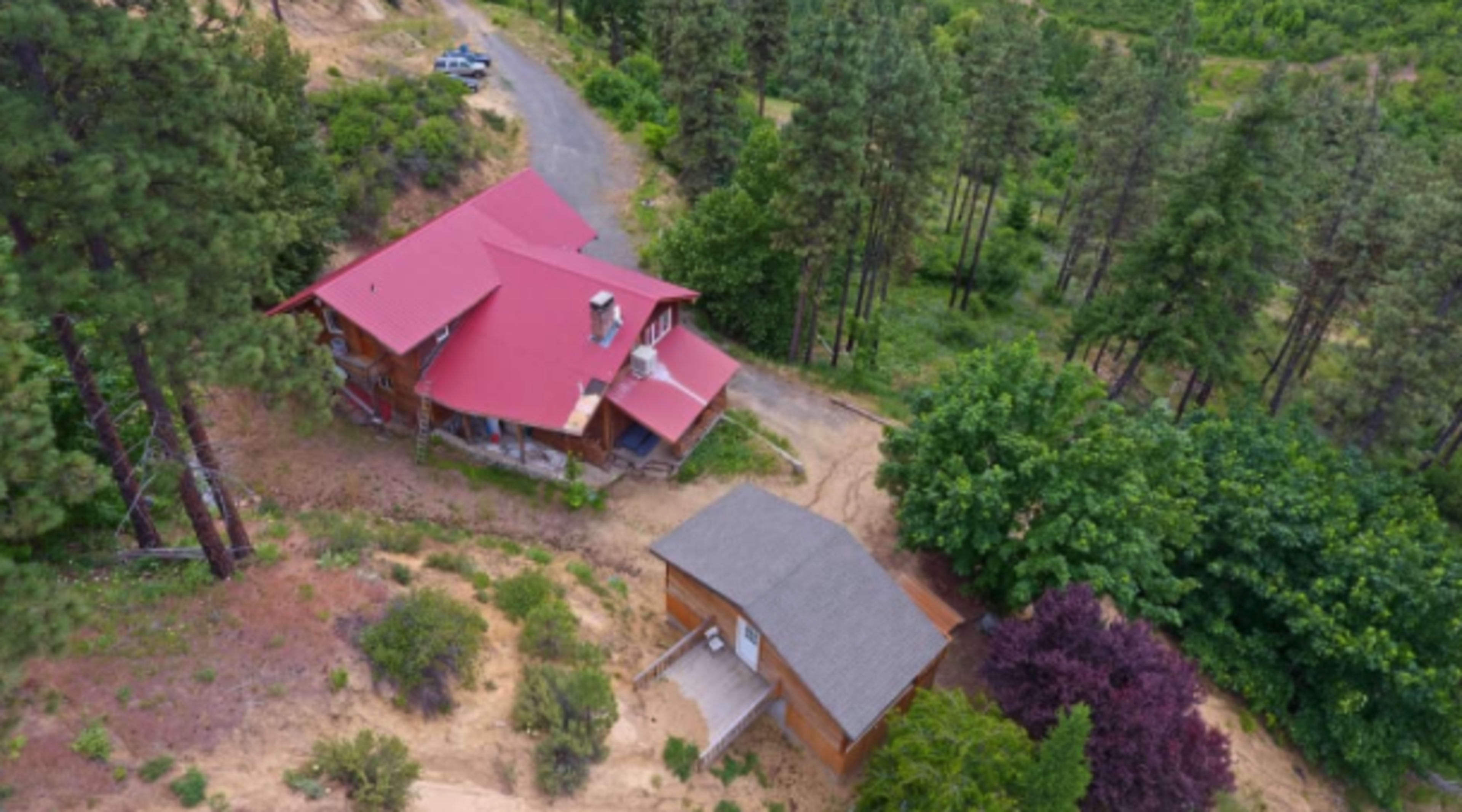 The image shows a wooden house with a red roof and an adjacent smaller building, surrounded by trees and a gravel road.