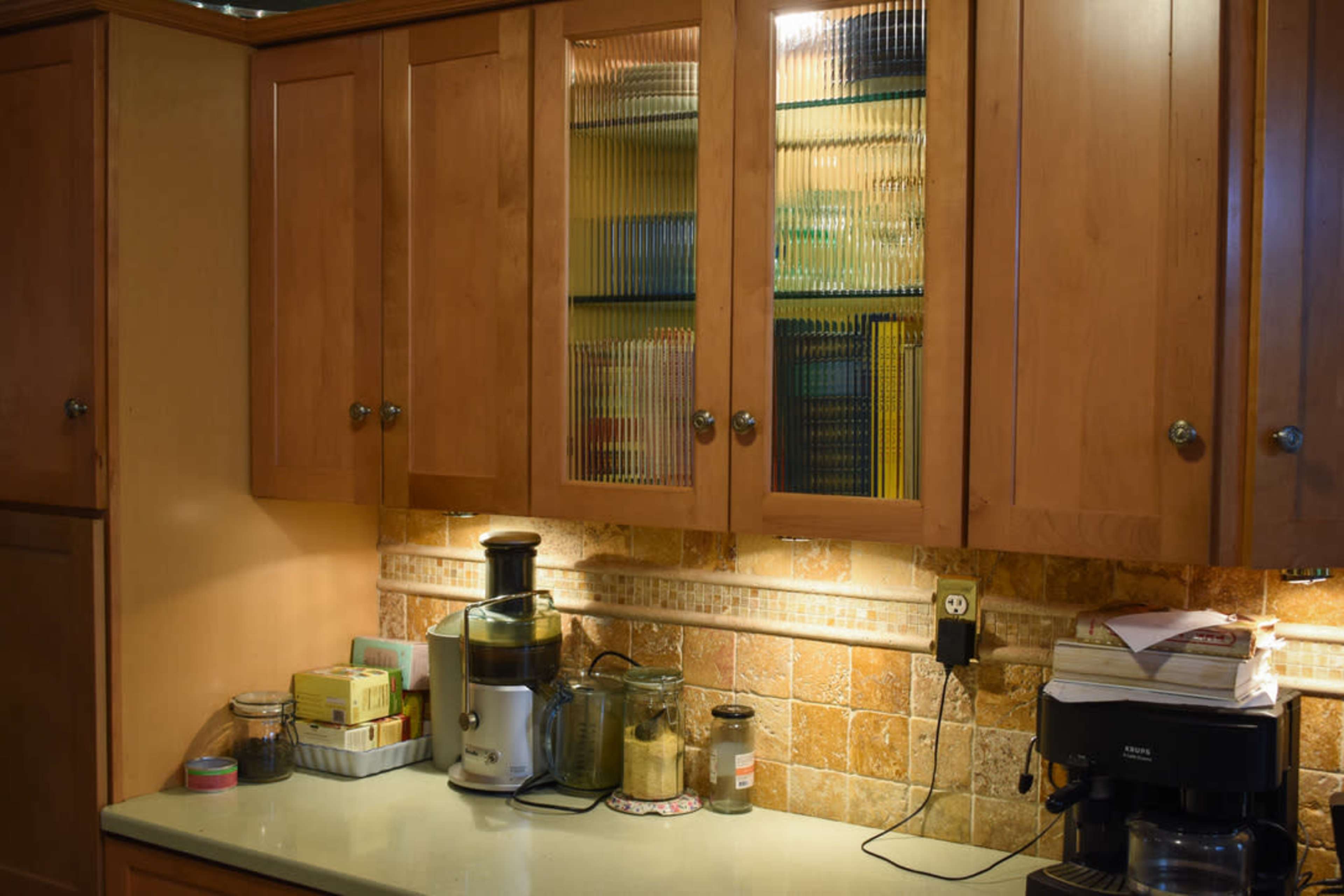 A kitchen features wooden cabinets with glass doors displaying colorful items, alongside a countertop with a juicer and various containers.