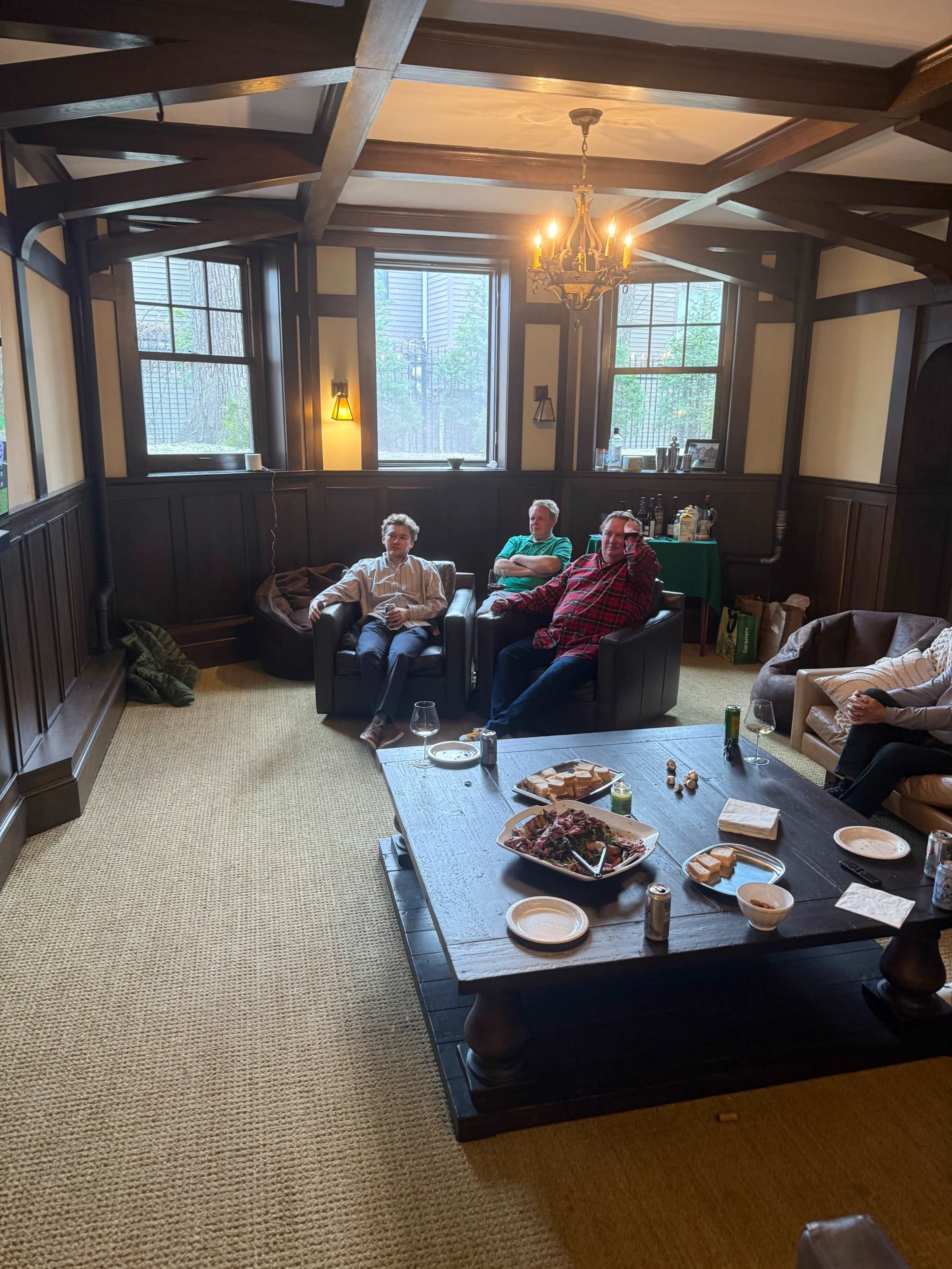 A group of four people is seated in a cozy living room with dark wooden paneling and a large table displaying food and drinks.