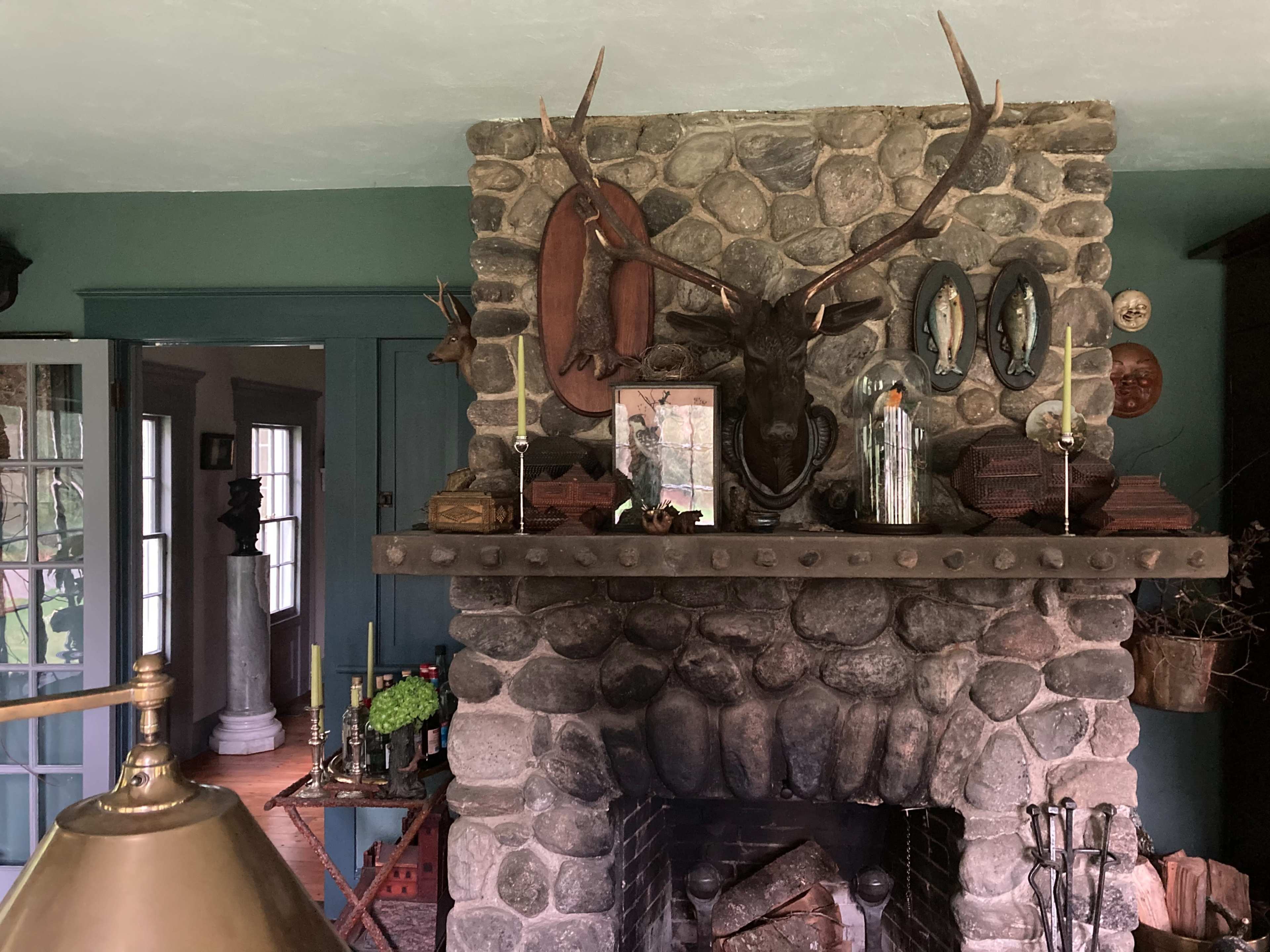 A stone fireplace with antlers mounted above a wooden mantel adorned with candles, decorative items, and framed pictures, set against a dark green wall.