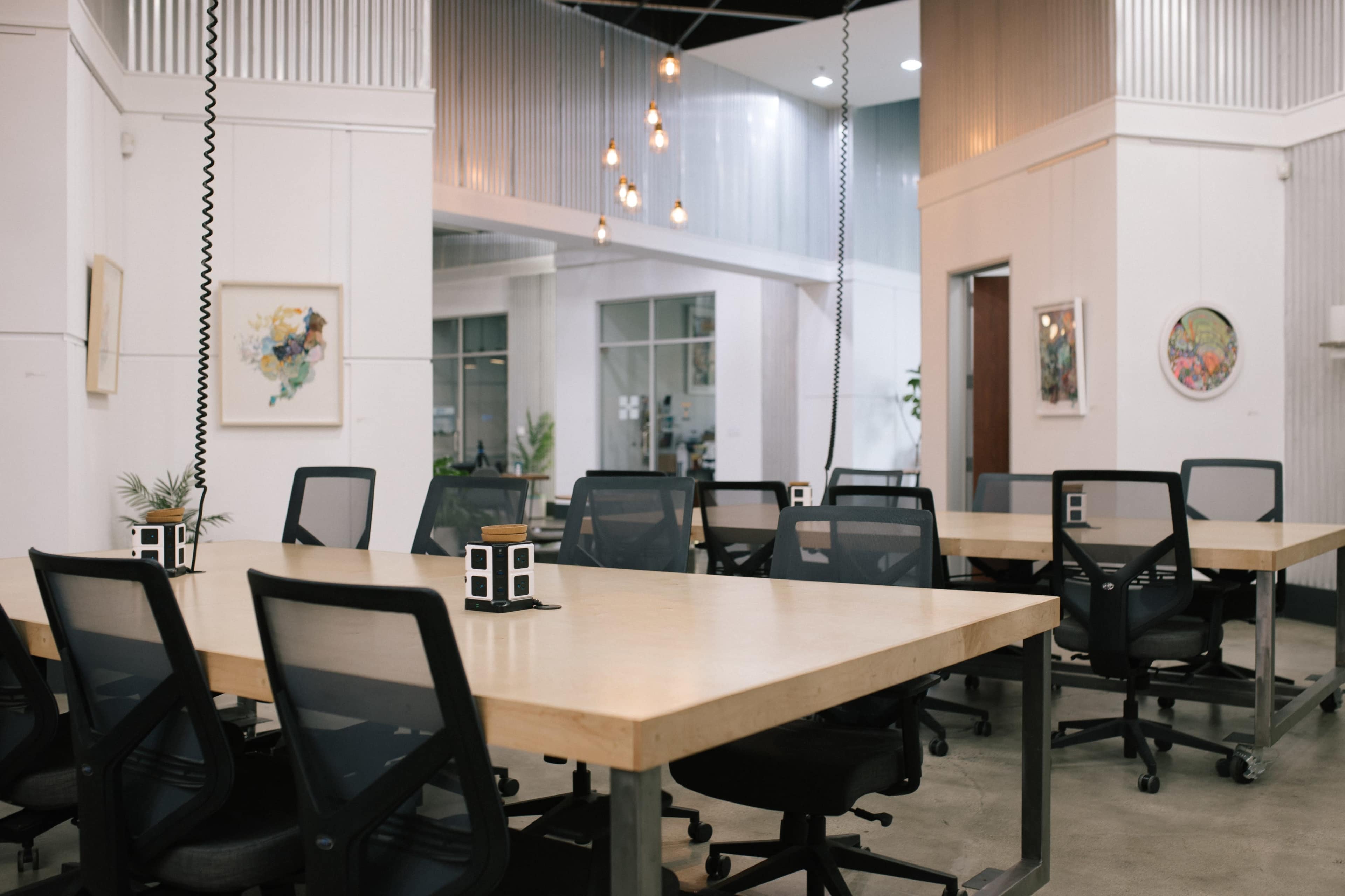 The image shows a modern office space with multiple large tables surrounded by ergonomically designed chairs, illuminated by hanging pendant lights.
