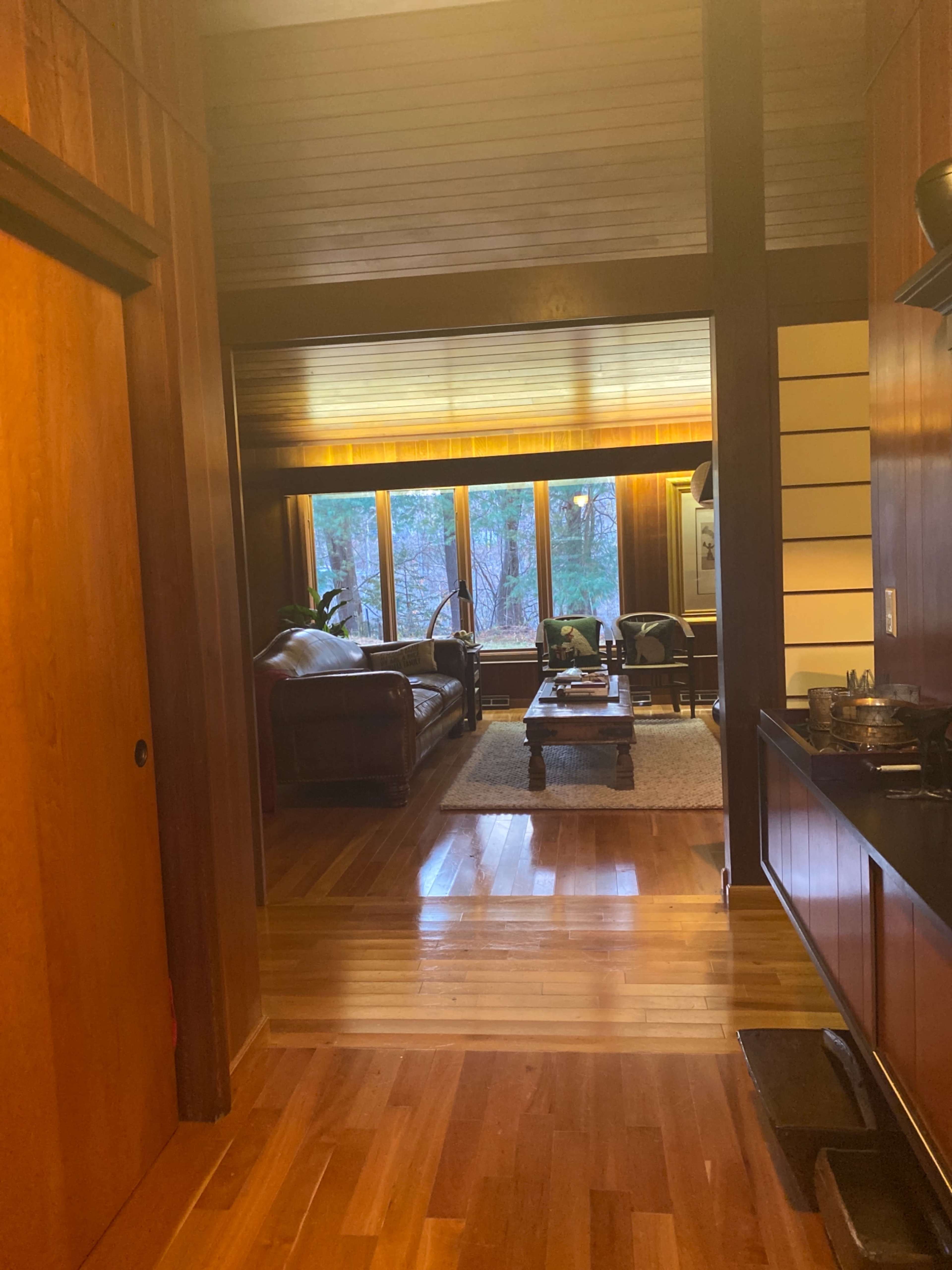 A wooden interior hallway leading to a living room with large windows overlooking a forested area.
