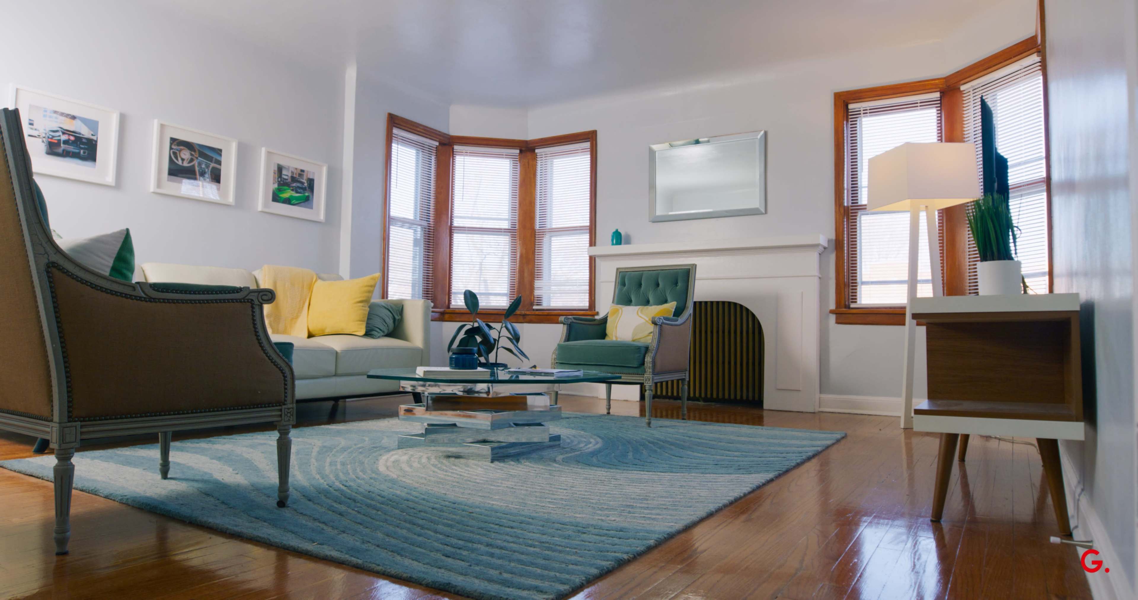 A cozy living room features large windows, a light-colored sofa with yellow and green accent pillows, and a coffee table on a circular patterned rug.