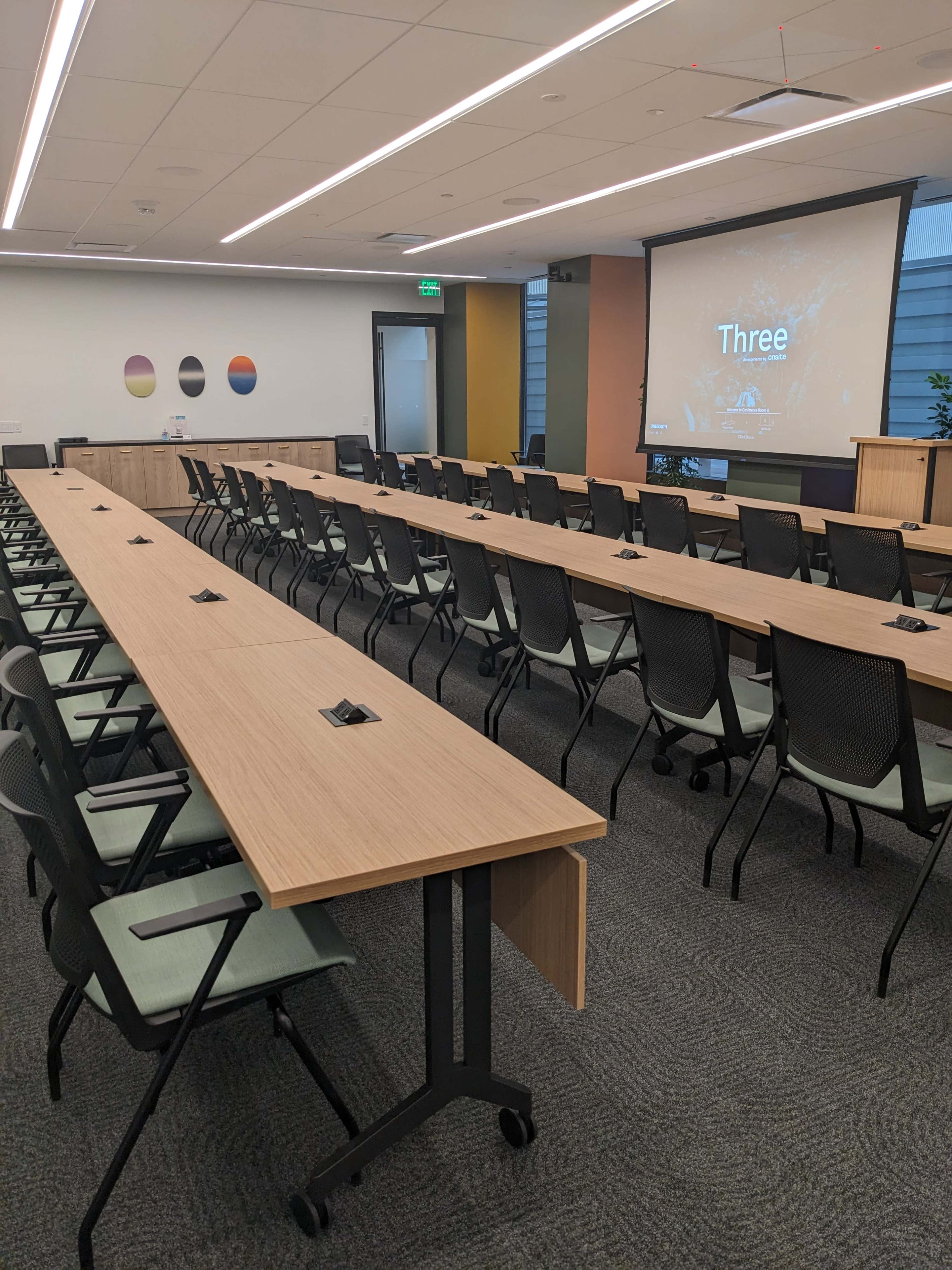 The image shows a conference room with multiple long tables arranged in rows, each equipped with chairs and a presentation screen displaying the word "Three."